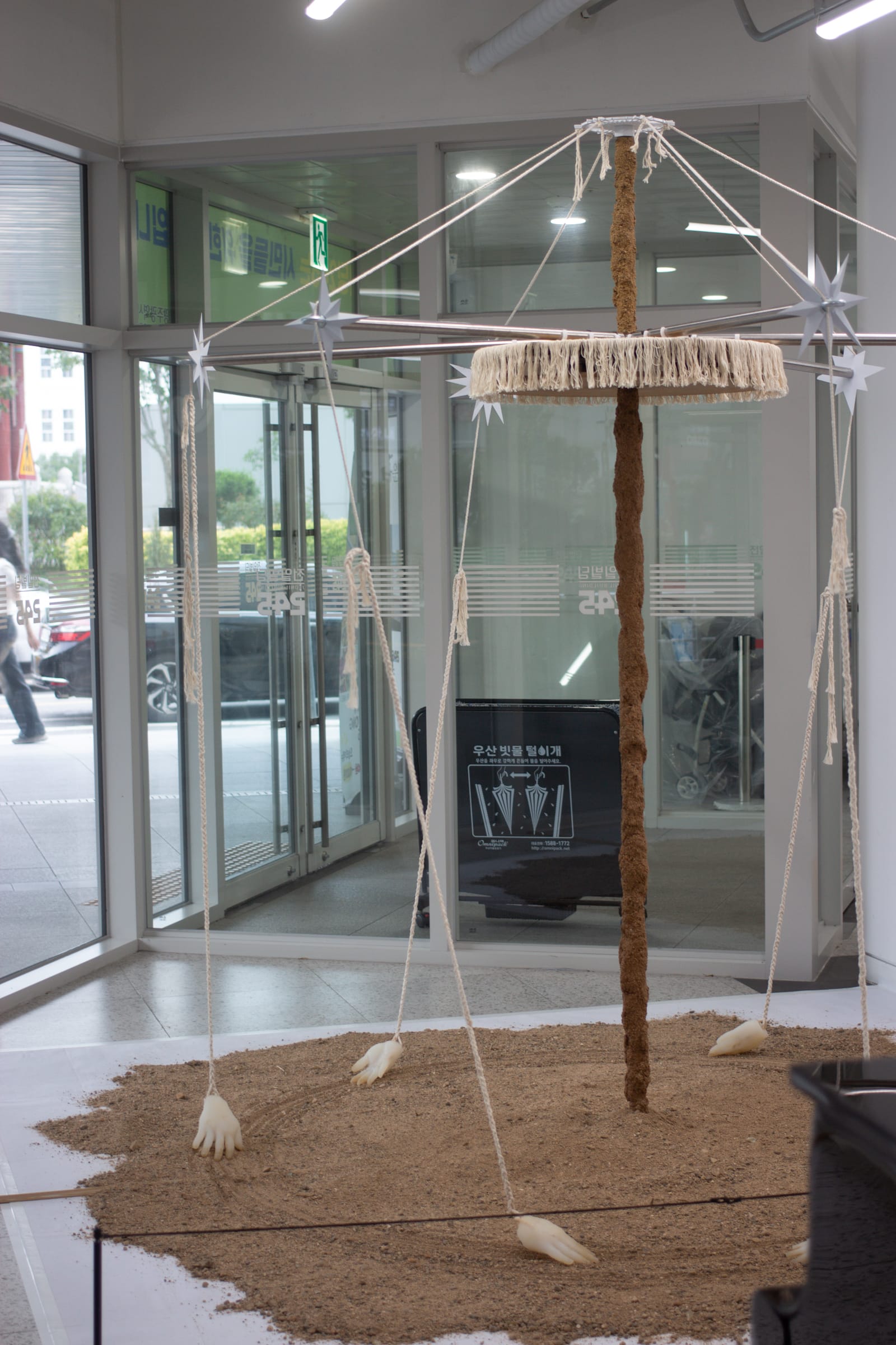 Art installation in the lobby of a building with glass walls. Six hand sculptures suspended by white rope appear to be dragged in a circle through sand, around a pole covered in sand and a rope-tasseled disc.