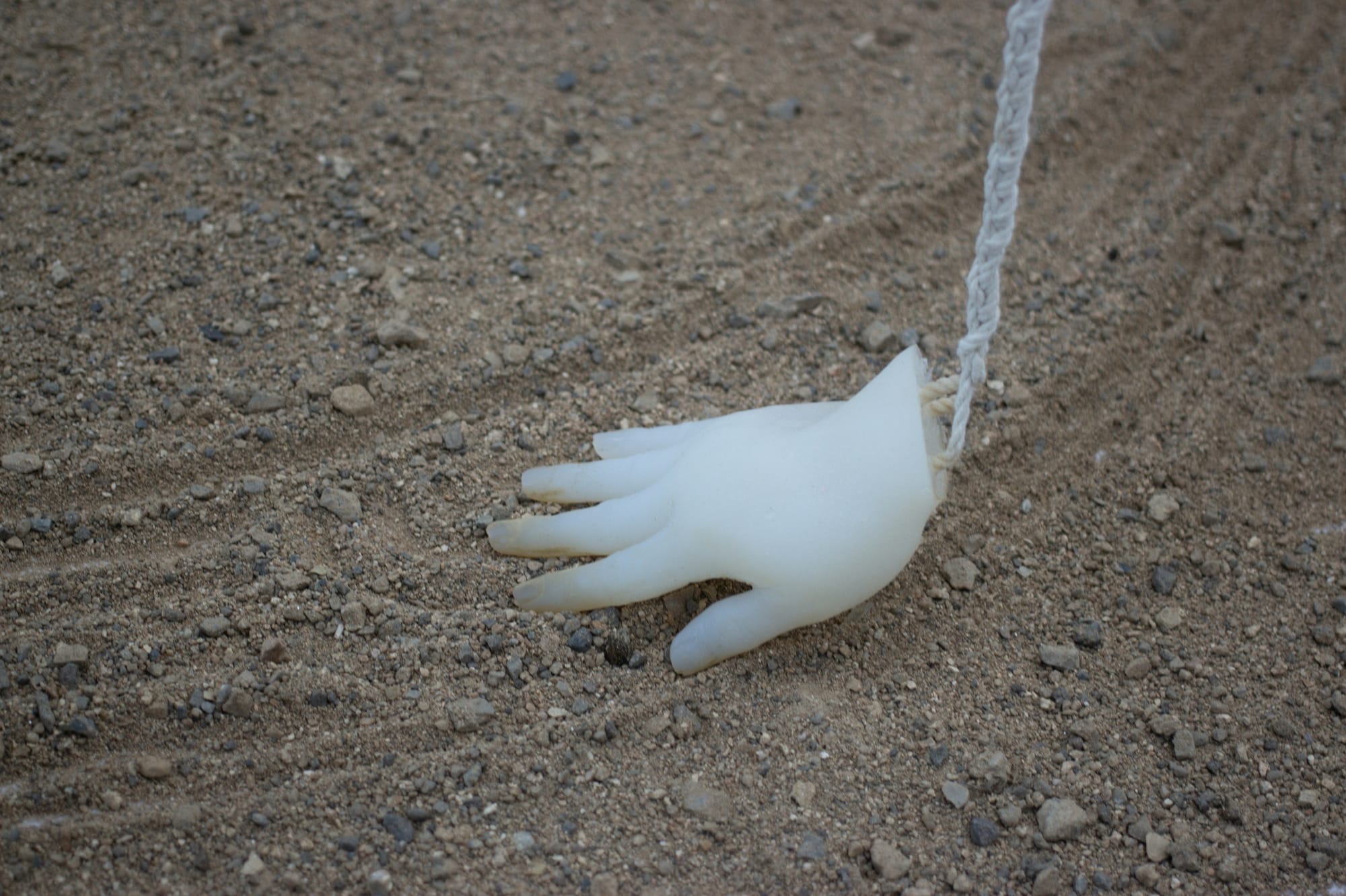 Close-up of translucent white hand sculpture suspended from white rope, making divots in the sand beneath it.