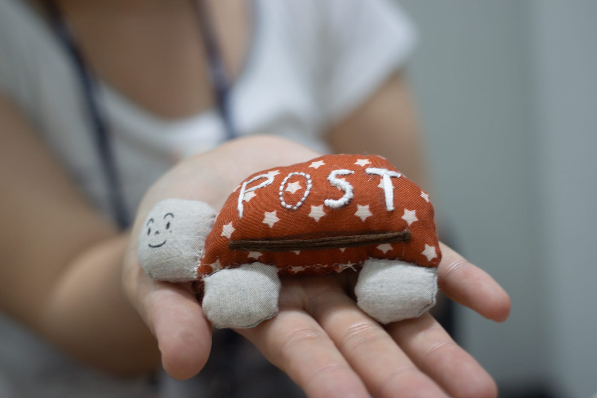 A hand holds out a fabric stuffed turtle with a smiling face, a brown mail slot, and the word "POST" on its shell.