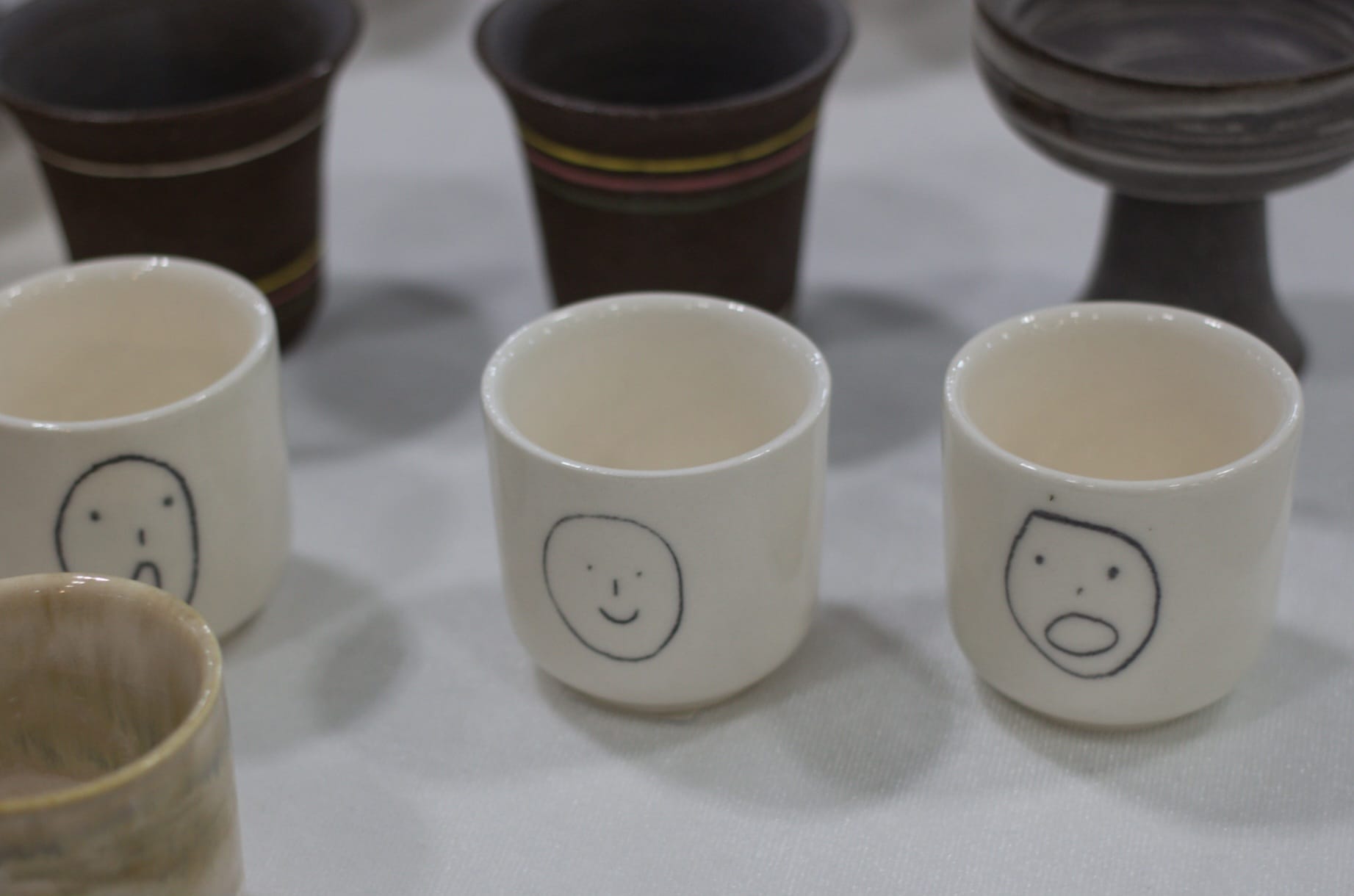 Three shot-glass-sized ceramic cups with simple line drawings of faces. Two are surprised, and one is smiling.