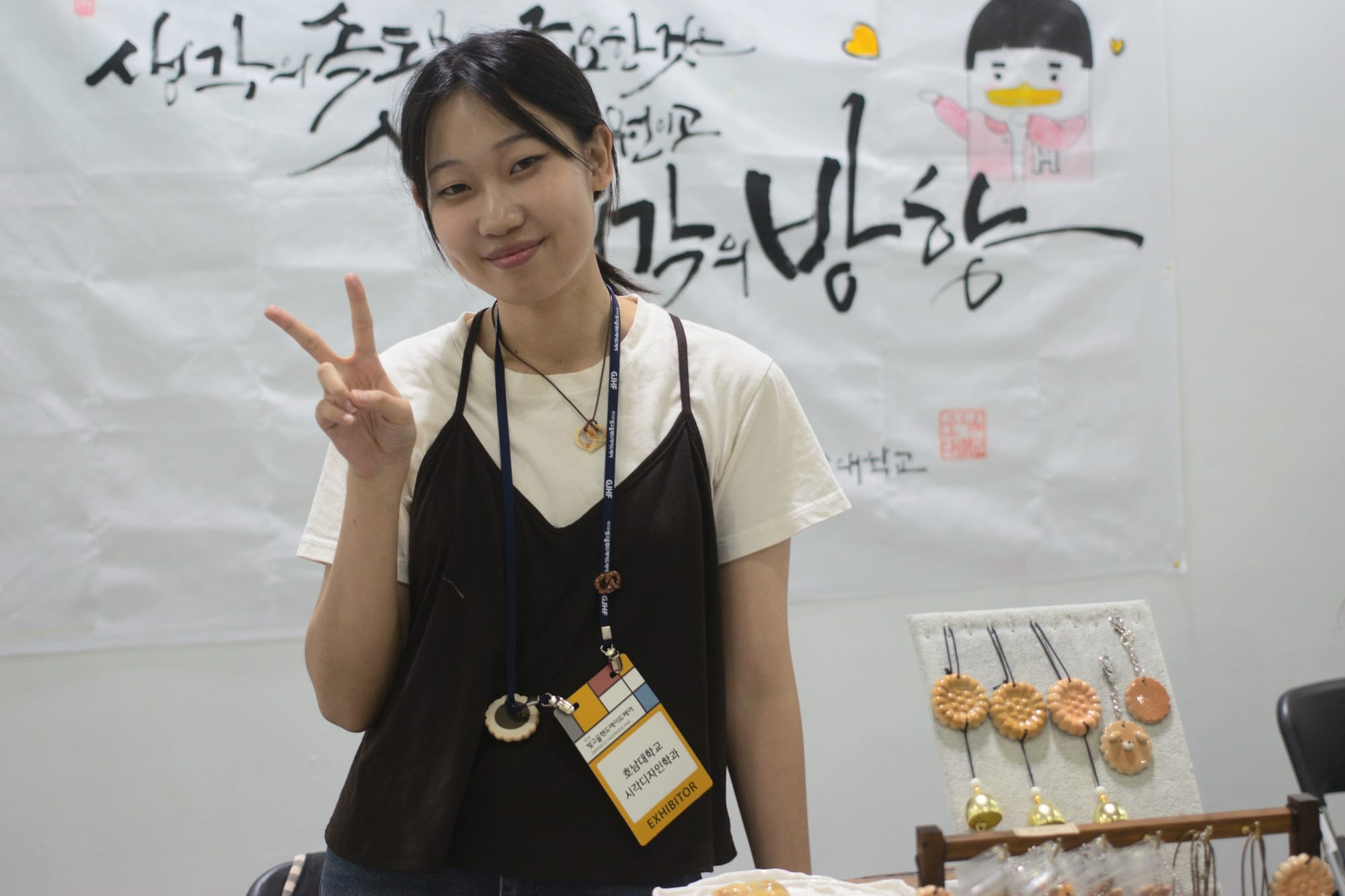 Cho Soyeon does a peace sign pose. She's standing at a booth. There's a banner behind her. In front are some ceramics in the shape of baked goods, some with bells.