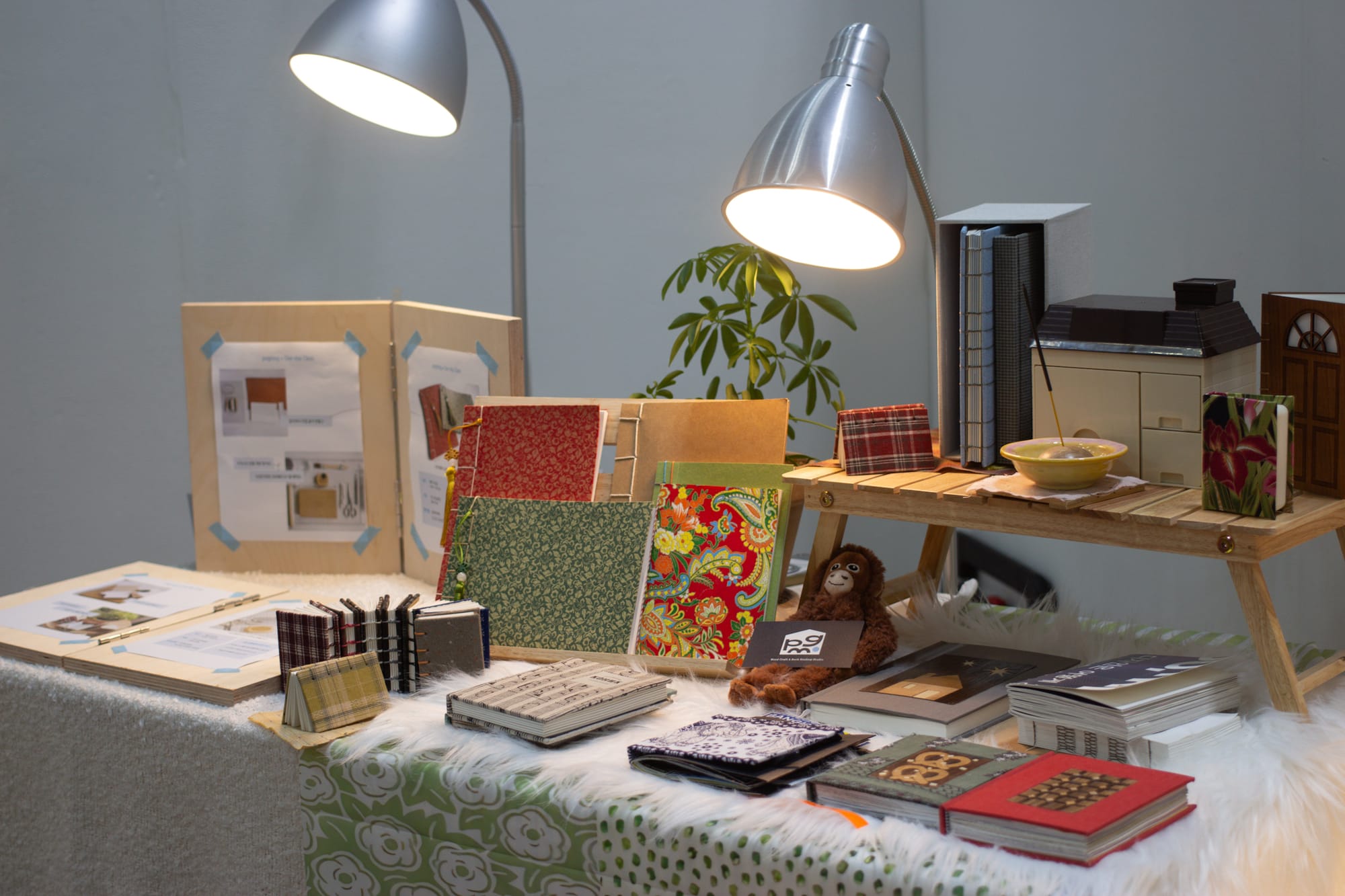 A display of miniature books with two metal lamps, a wooden riser, white faux-fur rug, and tablecloths of various patterns.