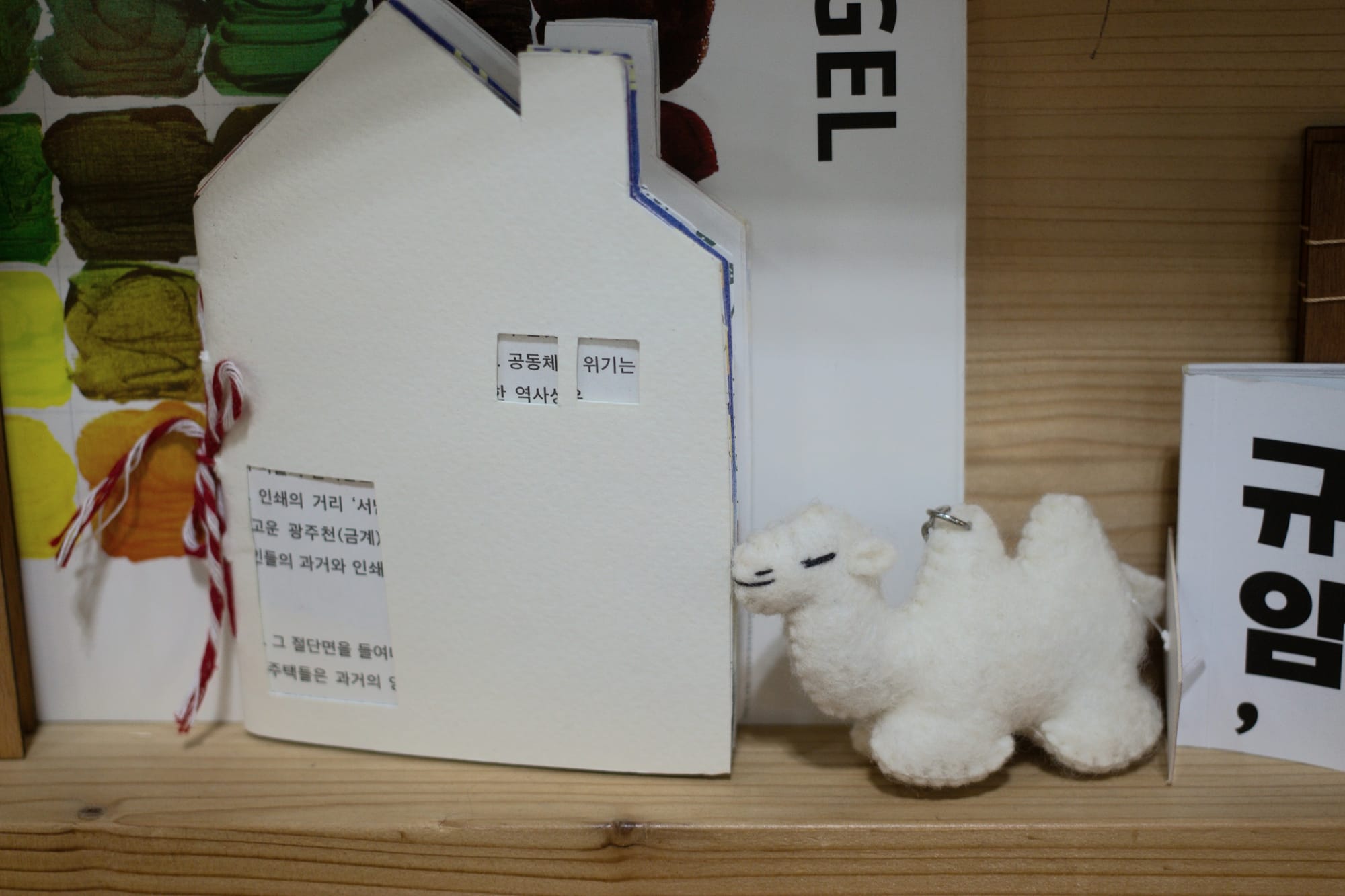 A thin hand-bound notebook cut in the shape of a house with a chimney. Cutouts on the cover are in the shape of a door and windows, revealing writing in Korean on the pages. Next to it is a fabric camel keychain.