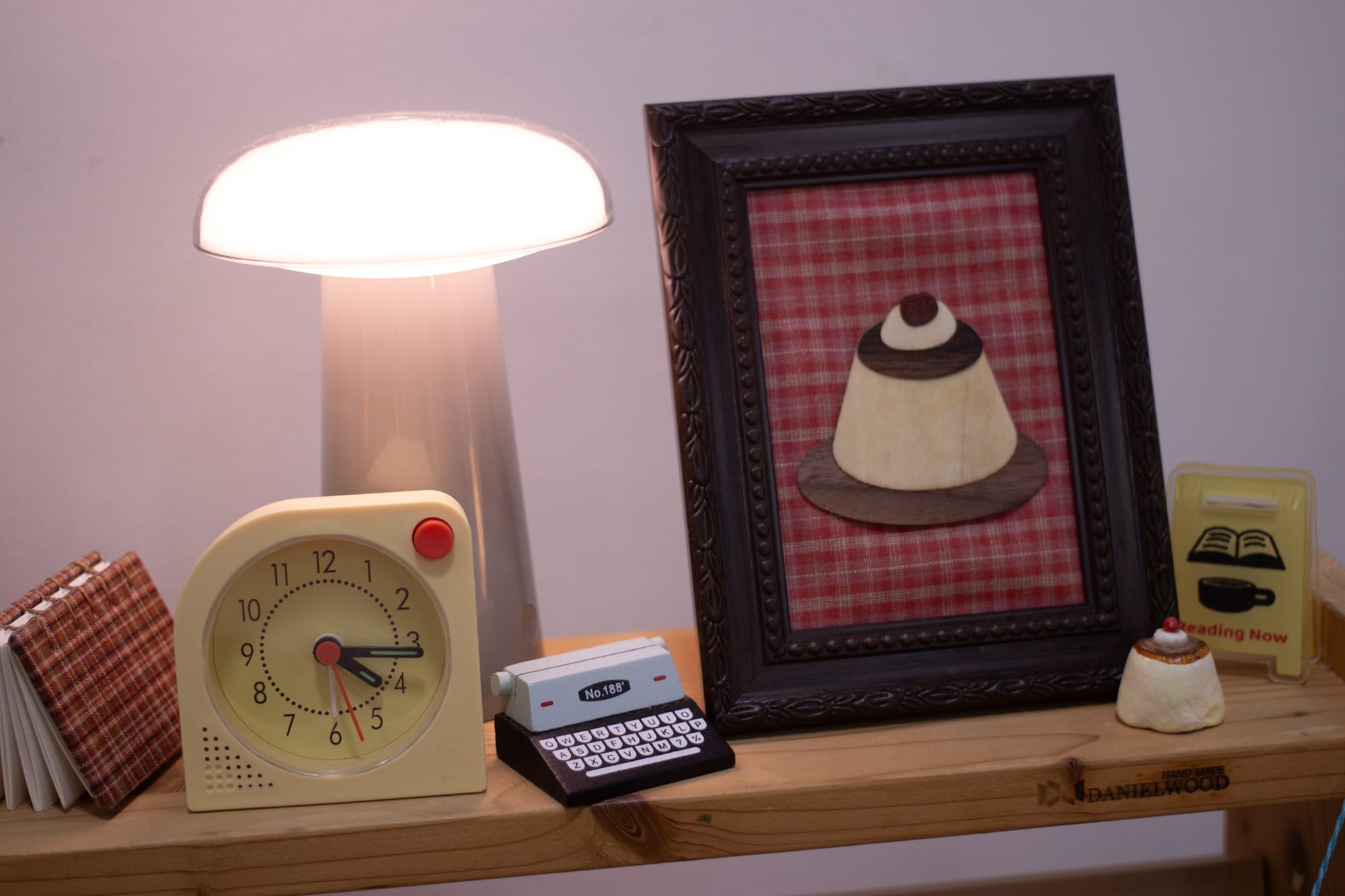 A small shelf with a tiny keyboard toy, a little retro analog clock, a mushroom-shaped lamp, a sign saying "Reading Now," a framed wooden collage of a cake, and an almost identical cake as a 3D sculpture.