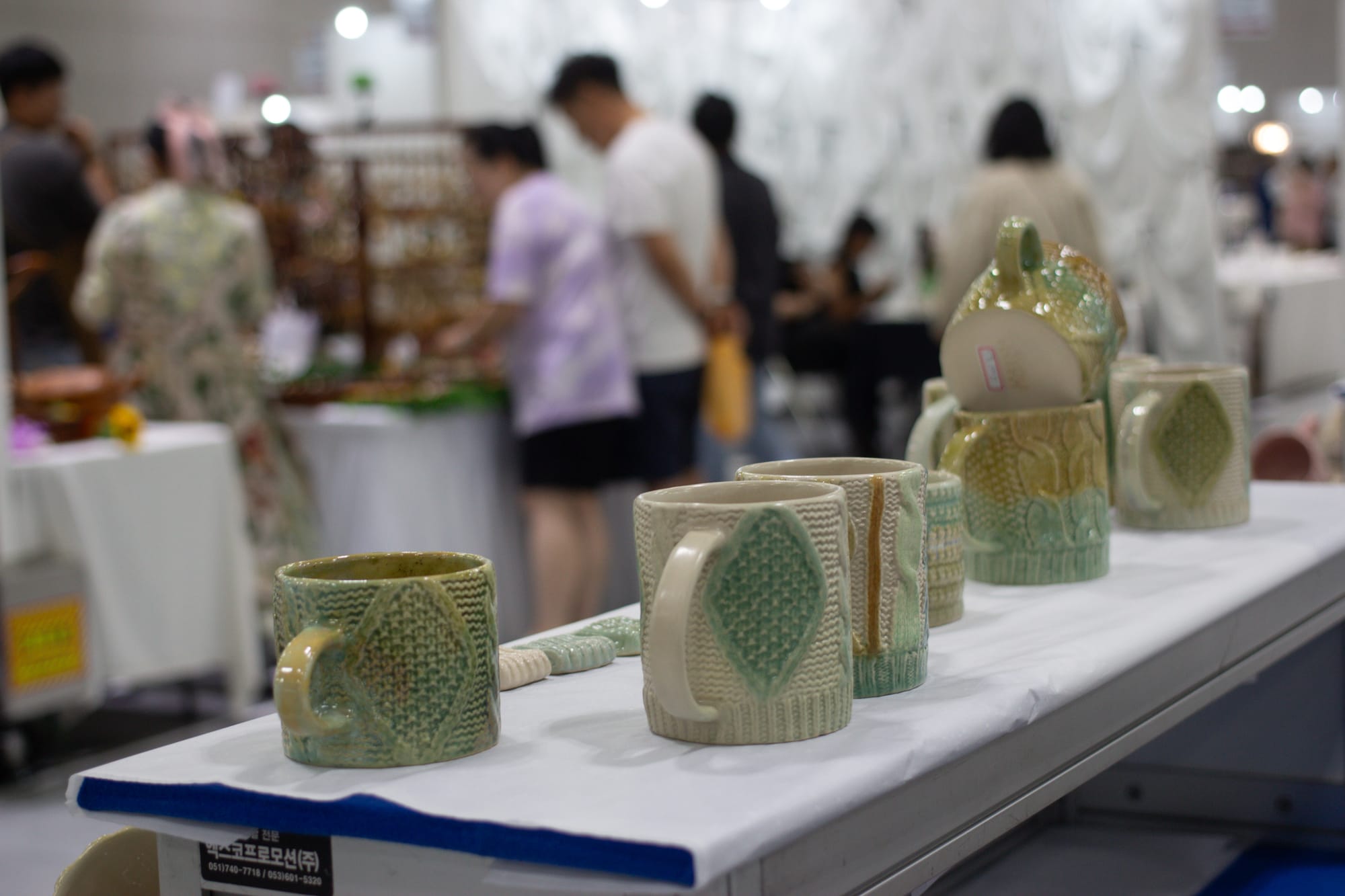 A display of green and beige ceramic mugs that look like knitted fabric. In the background are blurry people and other booths.