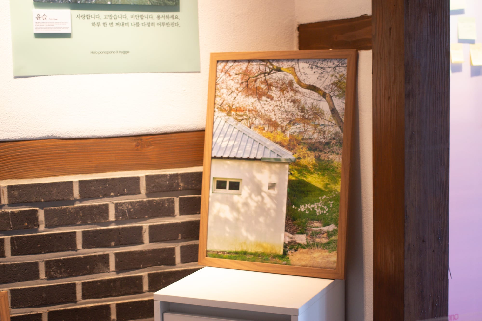 A large printed photograph in a wooden frame, resting in a corner. The photo is of a beige-colored building under some trees. The building exterior wall has dappled sunlight on it.