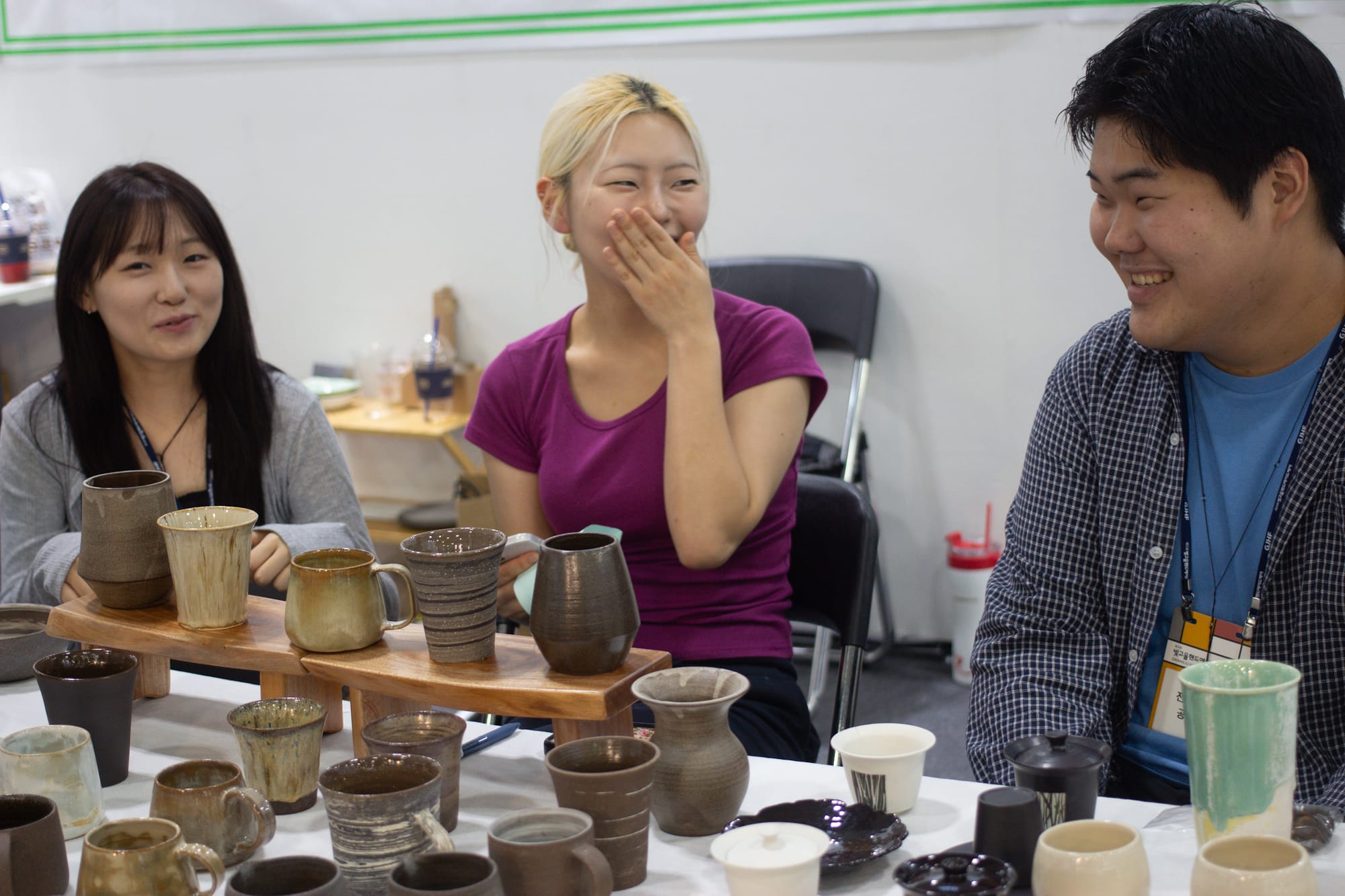 Three students sit, giggling, behind a display of ceramic mugs in shades of brown.