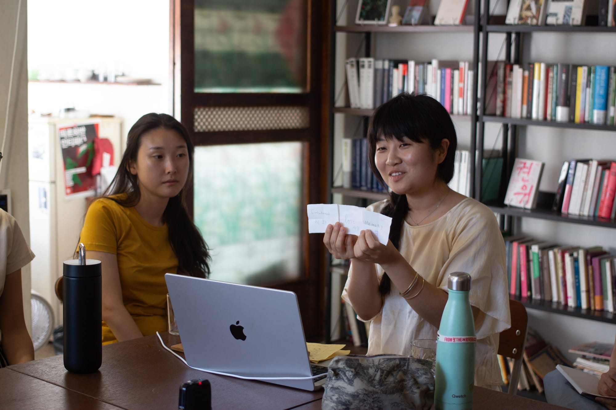 Haesu looks forward. Sue talks and holds up three white papers with words written on them. Behind is a bookshelf filled with books. On the table are some reusable water bottles and a laptop.