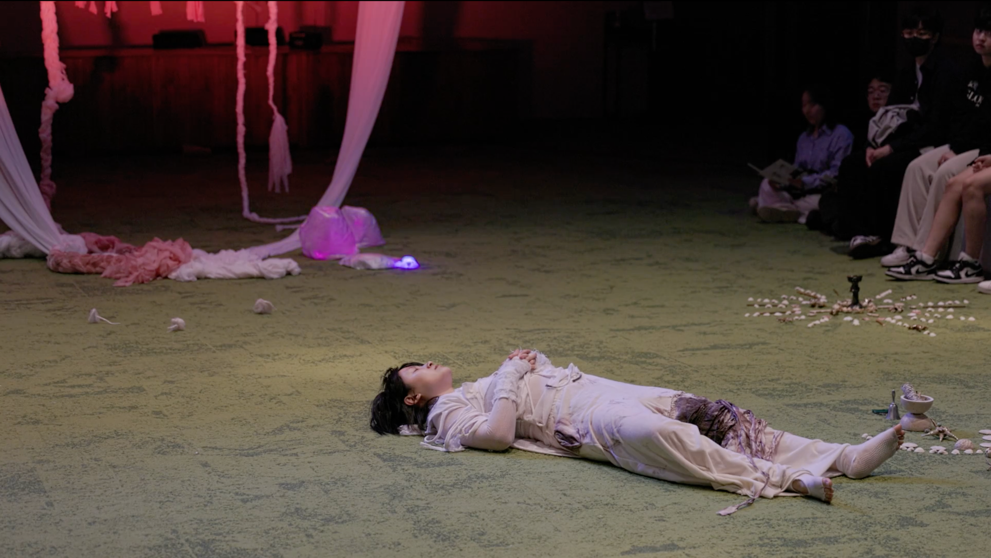 A play; some audience members are visible on the side. In the center, a person dressed in white rags lies on the floor. Also on the floor are geometrically arranged ceramic objects. White curtains are draped in the back.