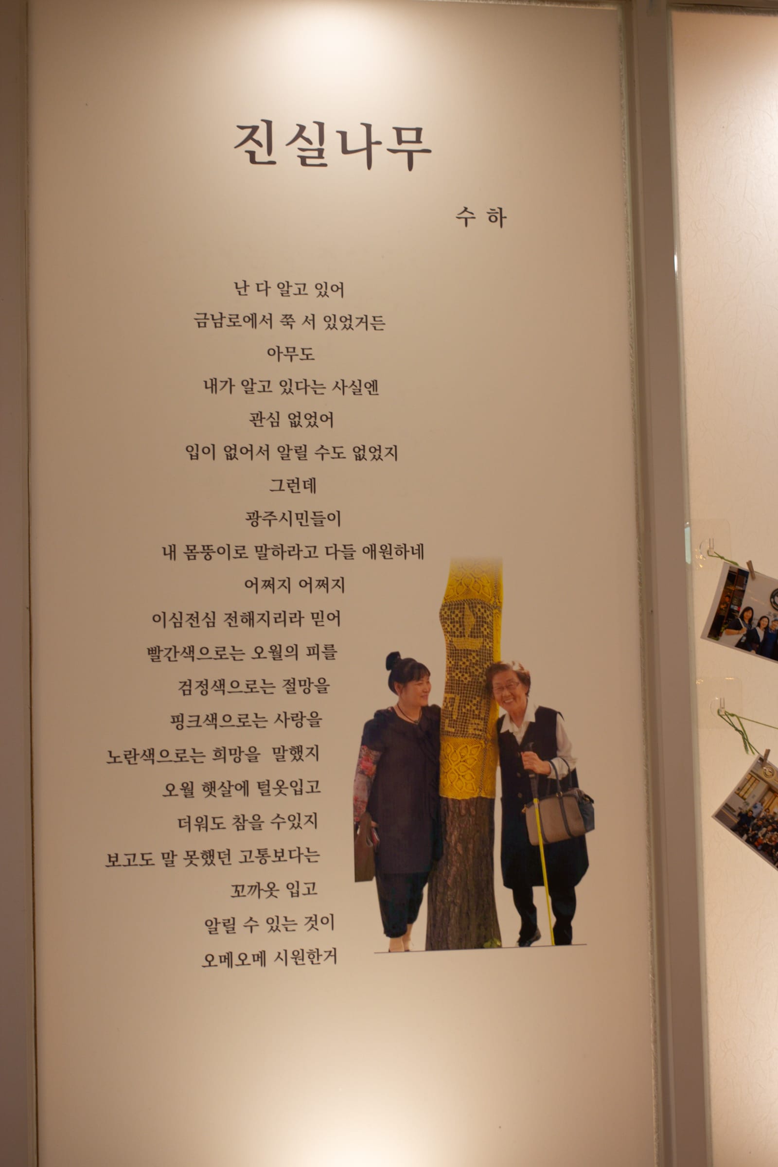 A poem printed in Korean on a wall, with a picture of Kim Suha and another woman, on either side of a tree wearing a crocheted yellow covering with a ship motif and "truth" in Korean.