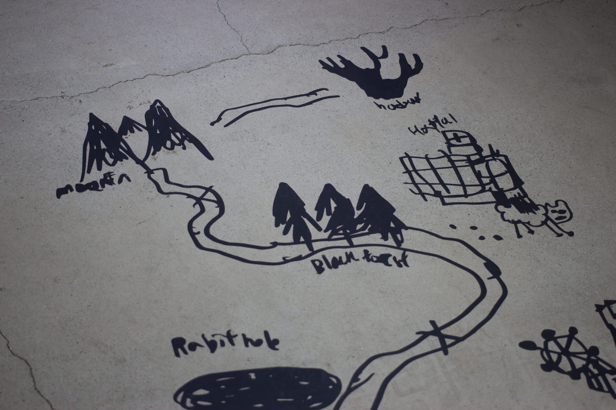Vinyl cutout art on a cracked concrete floor. It's a simple map through locations like "mountain," "Black forest," and "Rabit hole," each with a drawing. At "Hospital," there is a scribbled sheep.