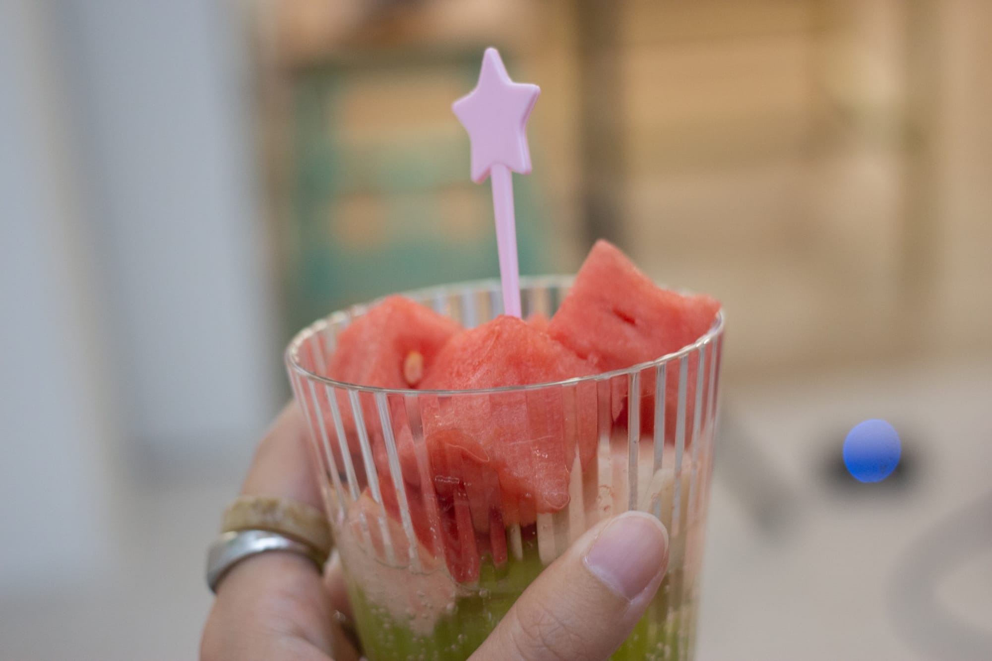 A hand with two rings holds a clear cup with watermelon cubes, peach pieces, and green grapes in a fizzy drink. There is a lavender plastic utensil inside in the shape of a star.