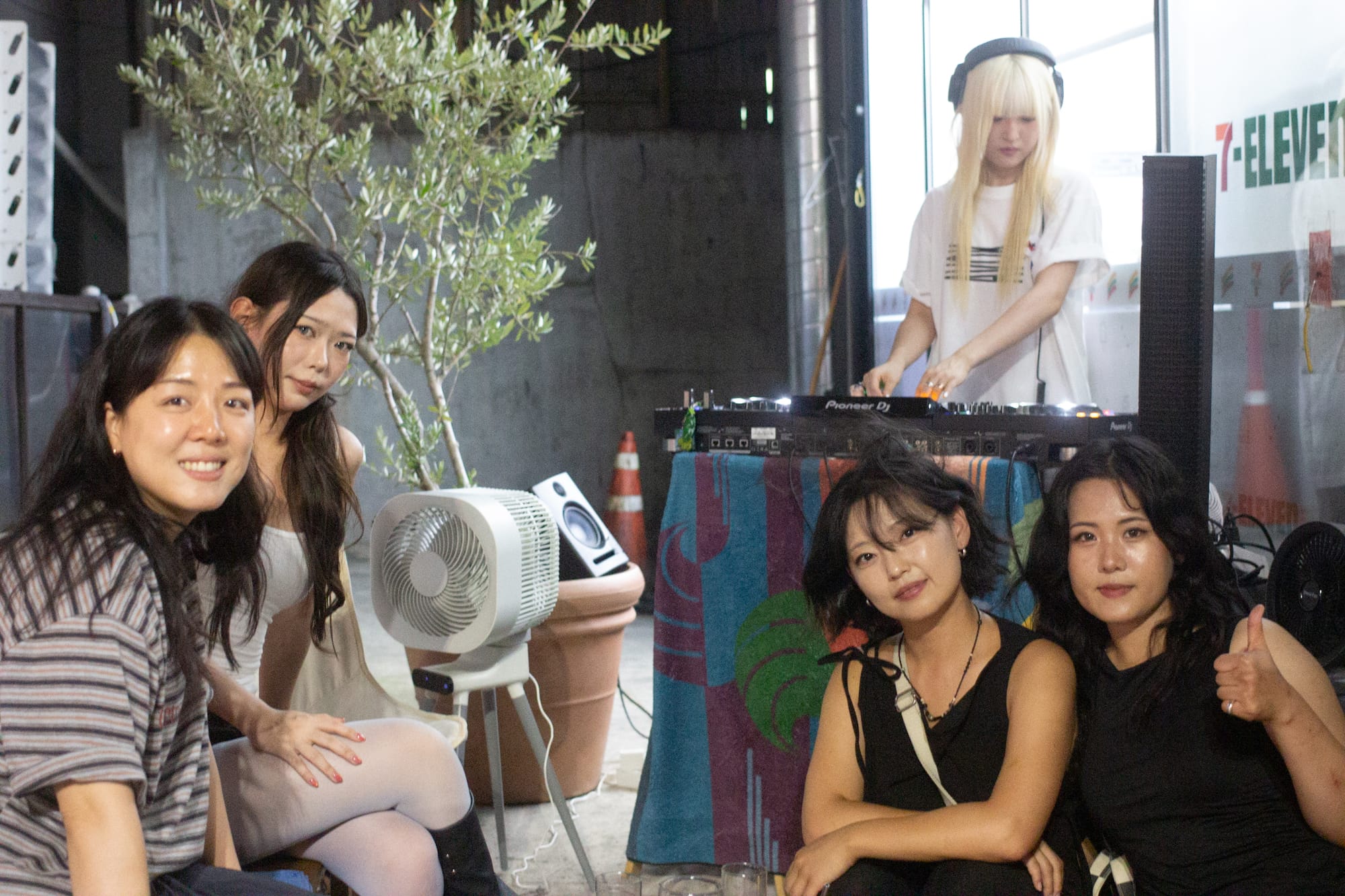 Four DJs sit in front of a table where another DJ is operating the decks. Behind is an open 7-Eleven, a fan, and a tree in a pot.