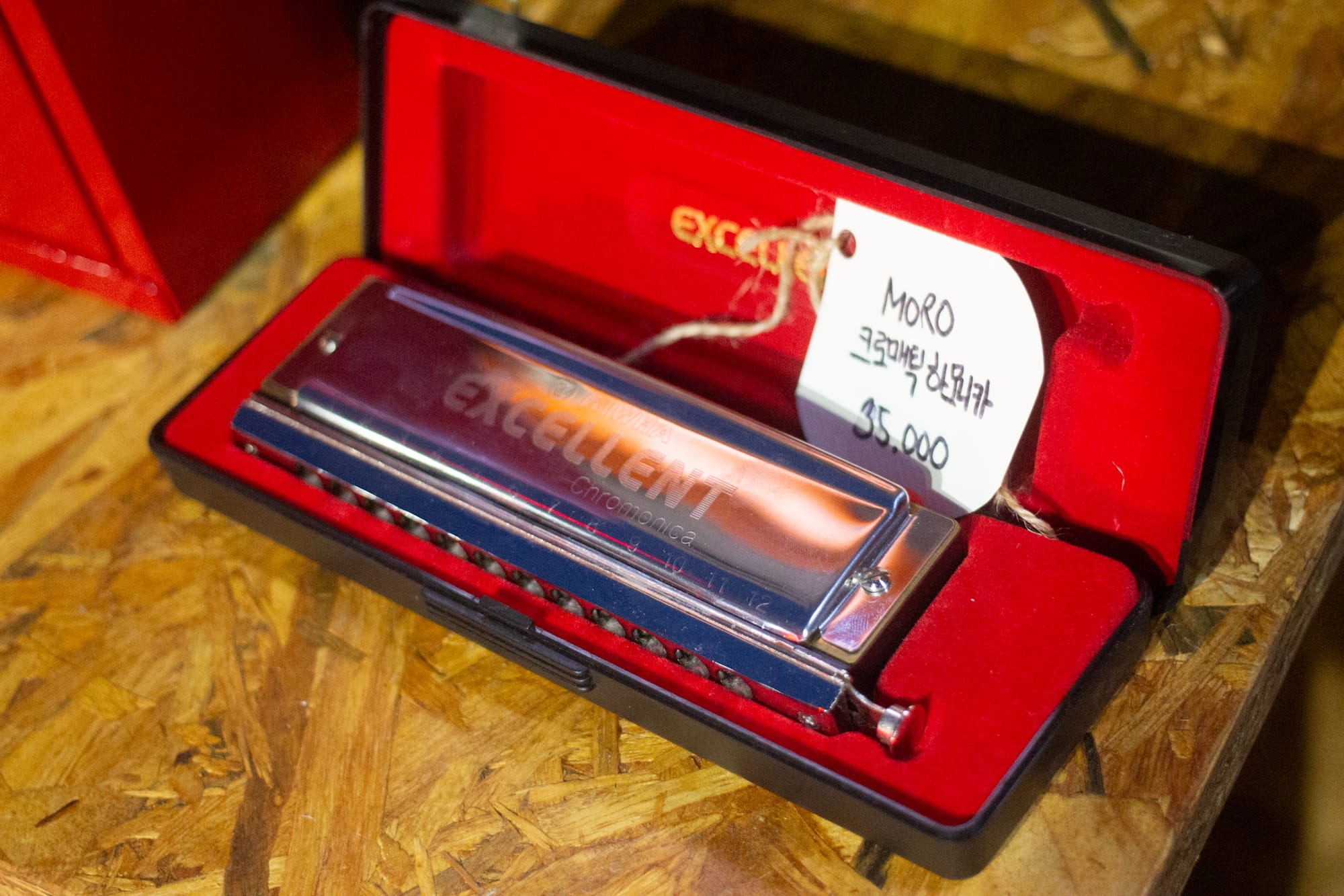 A metal harmonica bearing the words "EXCELLENT Chromonica" inside a red-lined case, with a handwritten tag labeling the item as DJ Moro's.