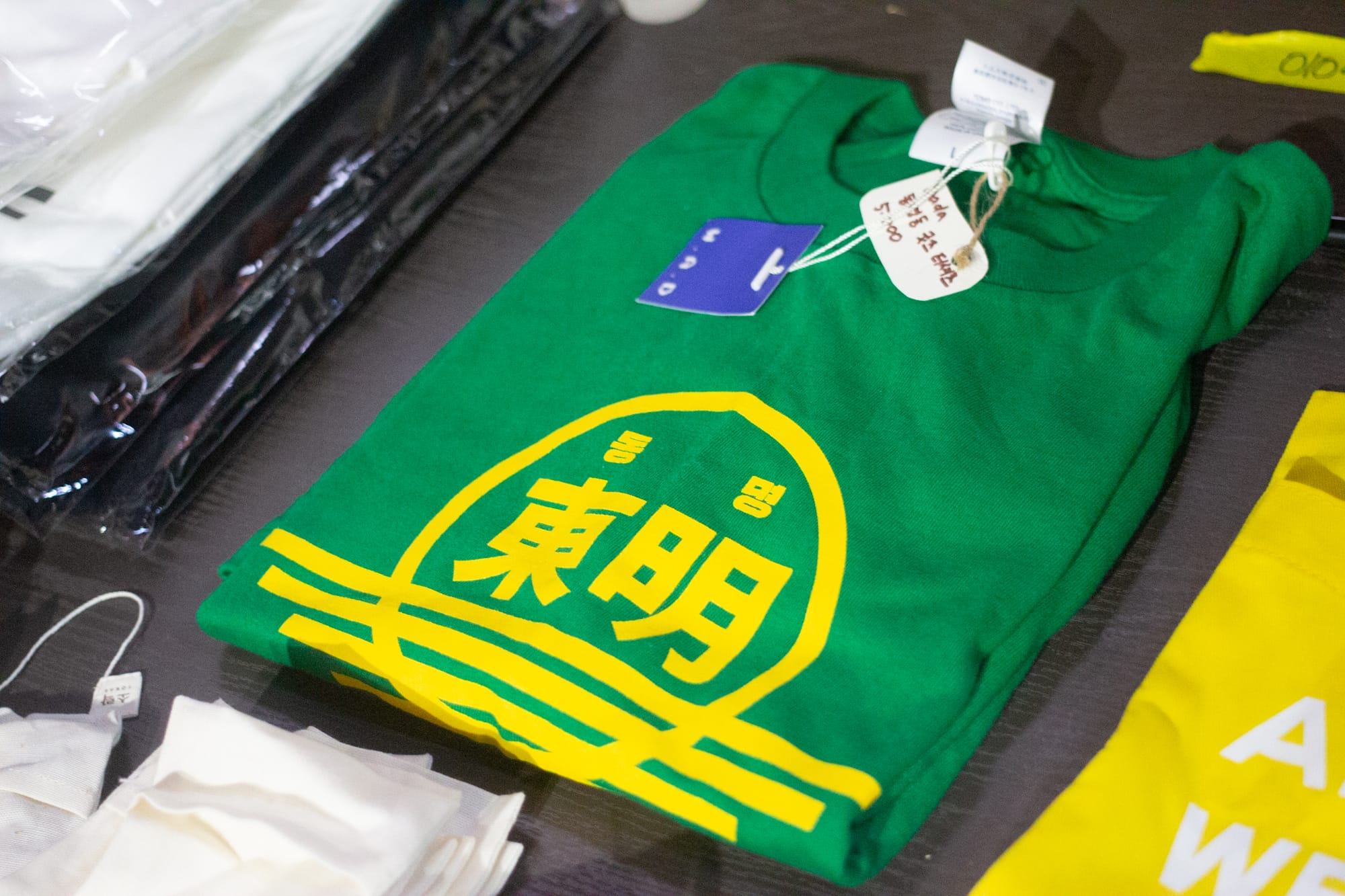 A folded green t-shirt with a yellow printed logo saying Dongmyeong in Korean letters and, below, Chinese characters saying "East" and "bright."