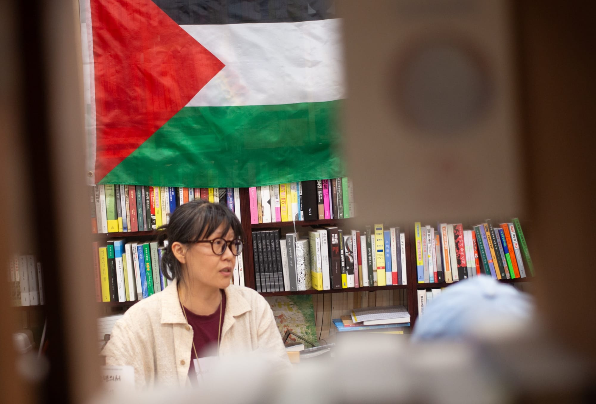 임인자 (Inza Lim) sits in front of filled bookshelves. Above her head is a Palestinian flag. The photo is taken through a bookshelf gap.