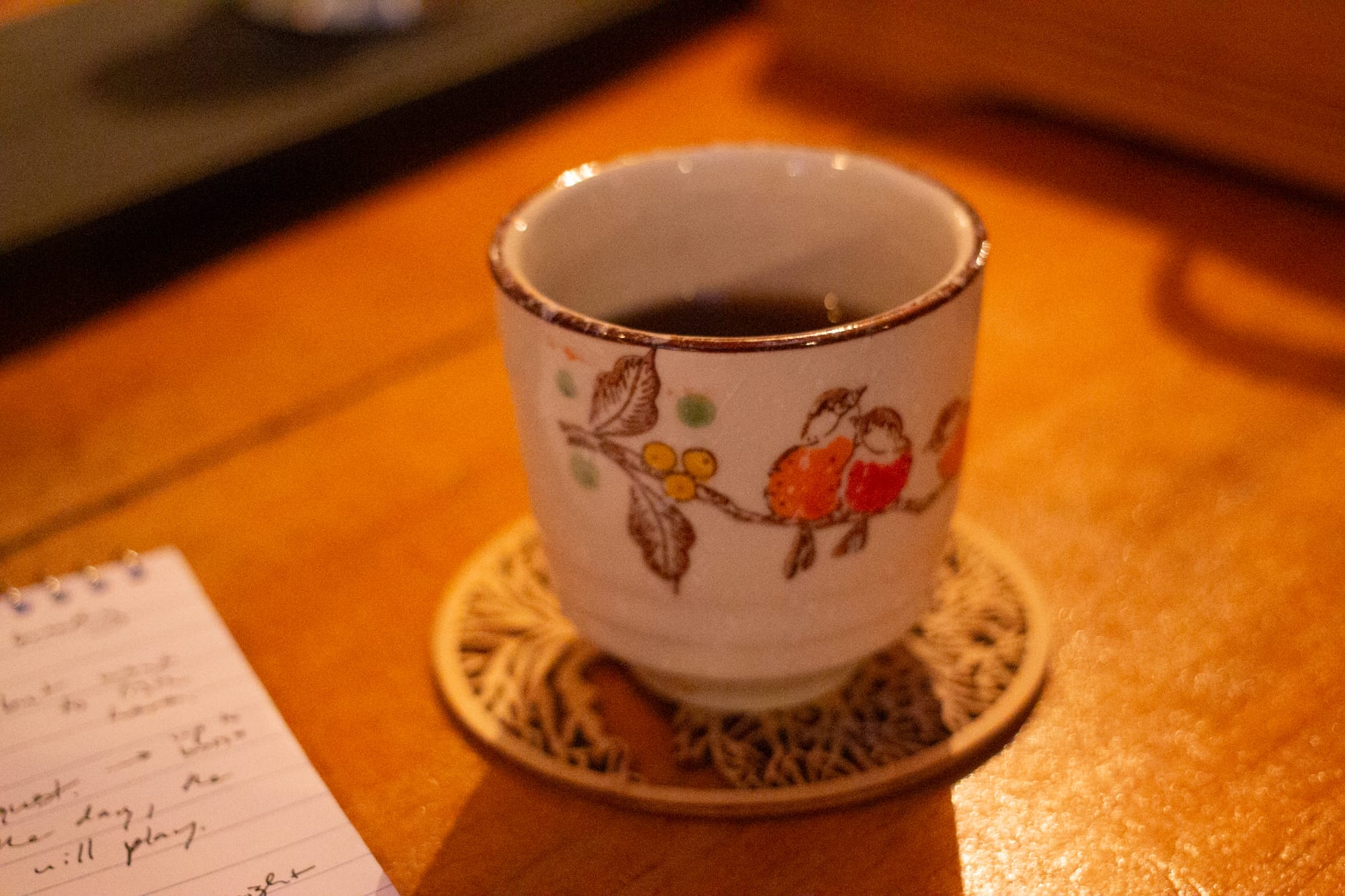 A teacup depicting two birds on a branch. A third bird can be seen slightly to the side. There is a dark liquid inside. The cup is on a wooden cutout coaster, and there is a corner of a notebook in the photo.