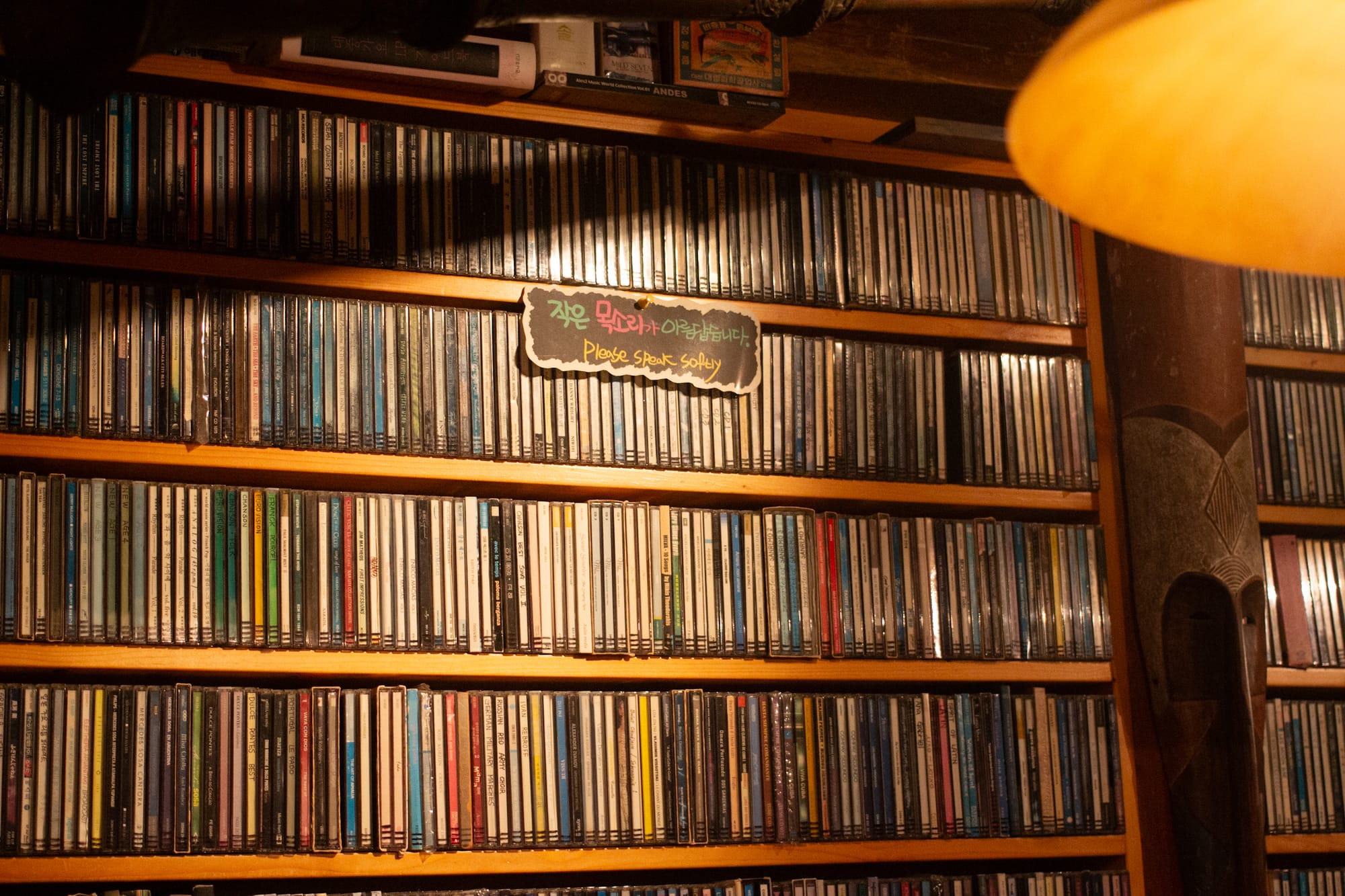 Light shines on shelves packed with CDs. A sign says "Please speak softly" in English and Korean.