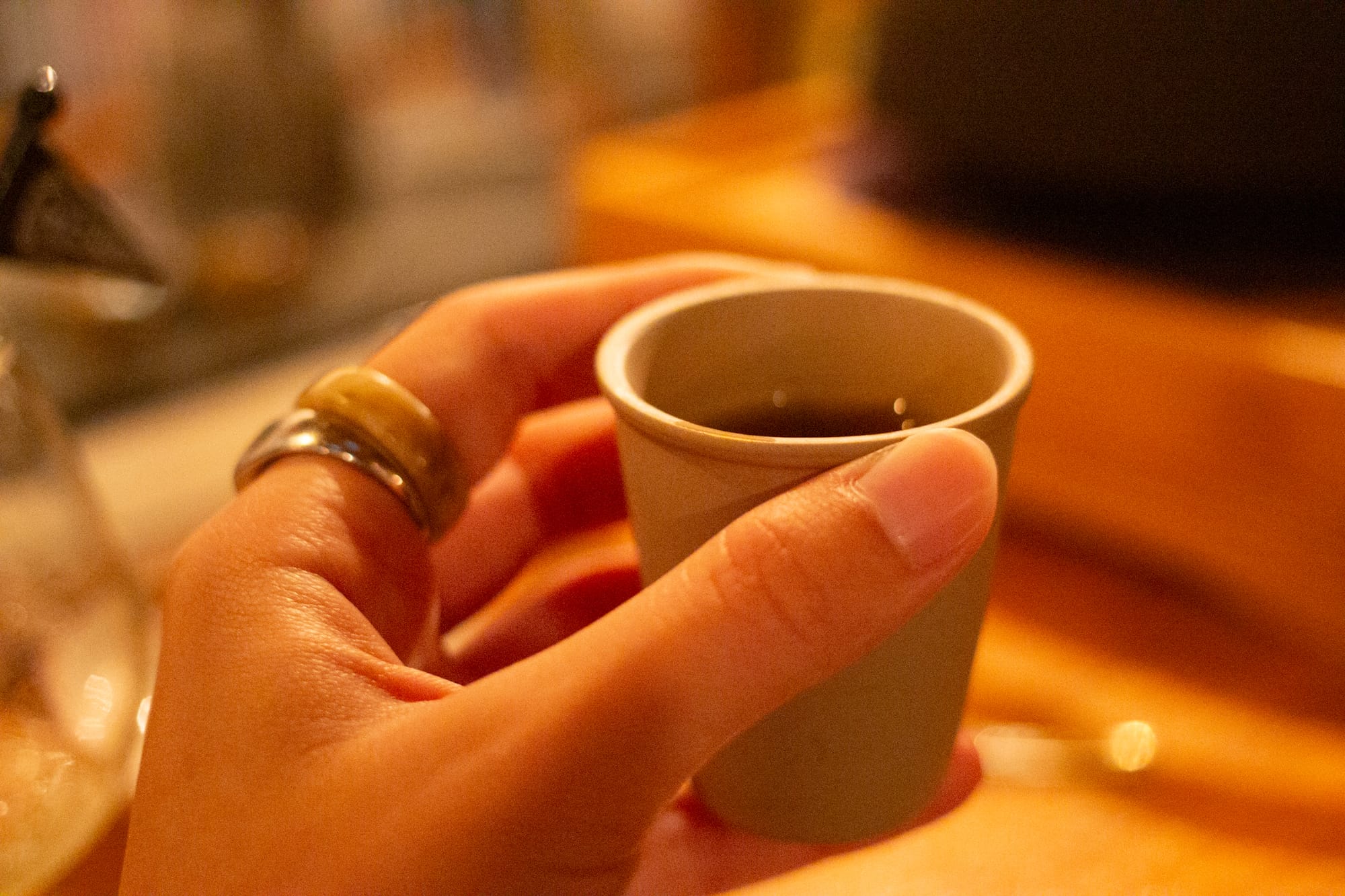A hand holds a tiny cup of tea in a warm-toned bar setting.