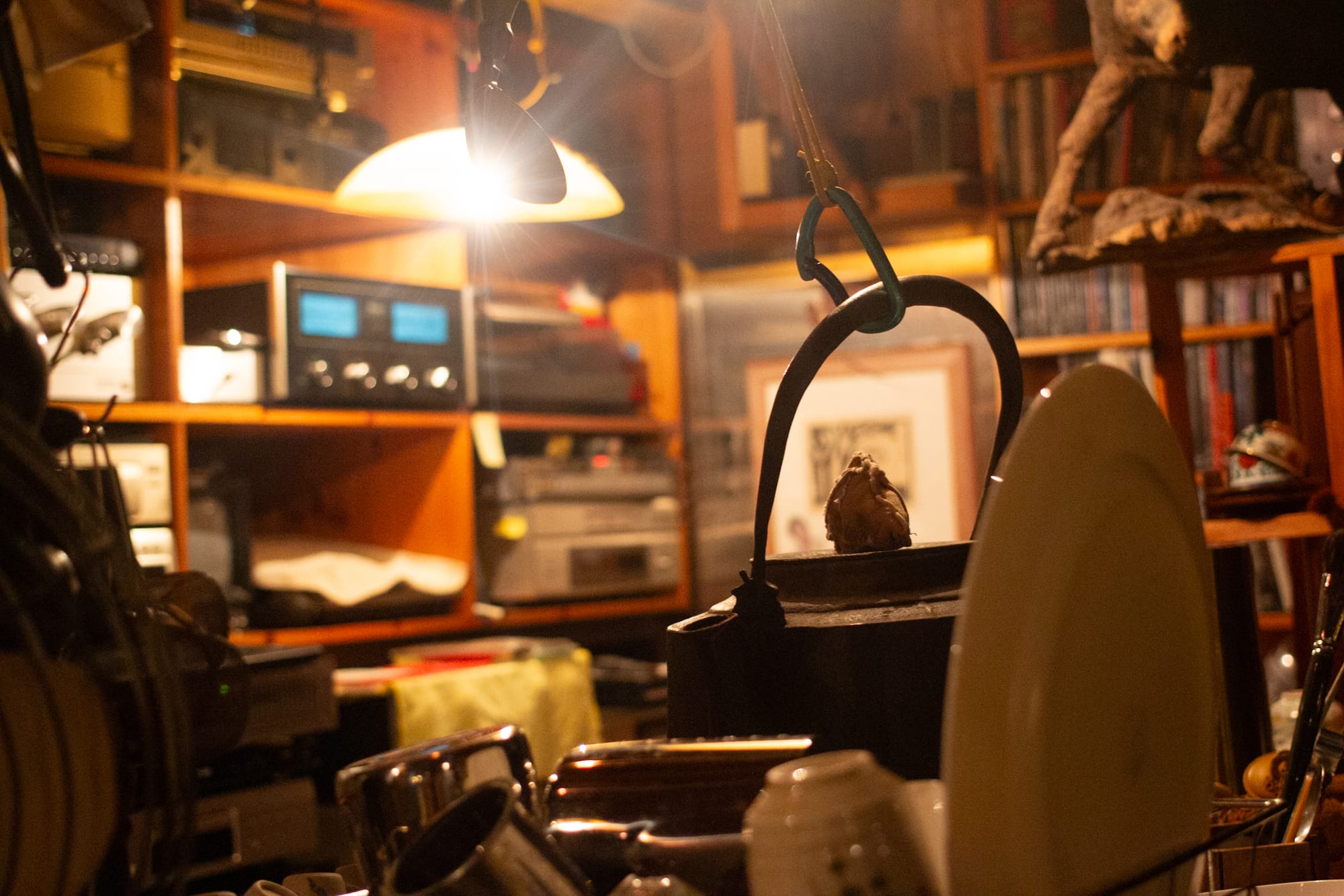 A domed lamp shines over a behind-bar interior filled with items on wooden shelves. A carabiner is attached to the handle of a black kettle. In the foreground is a plate and upside-down cup.