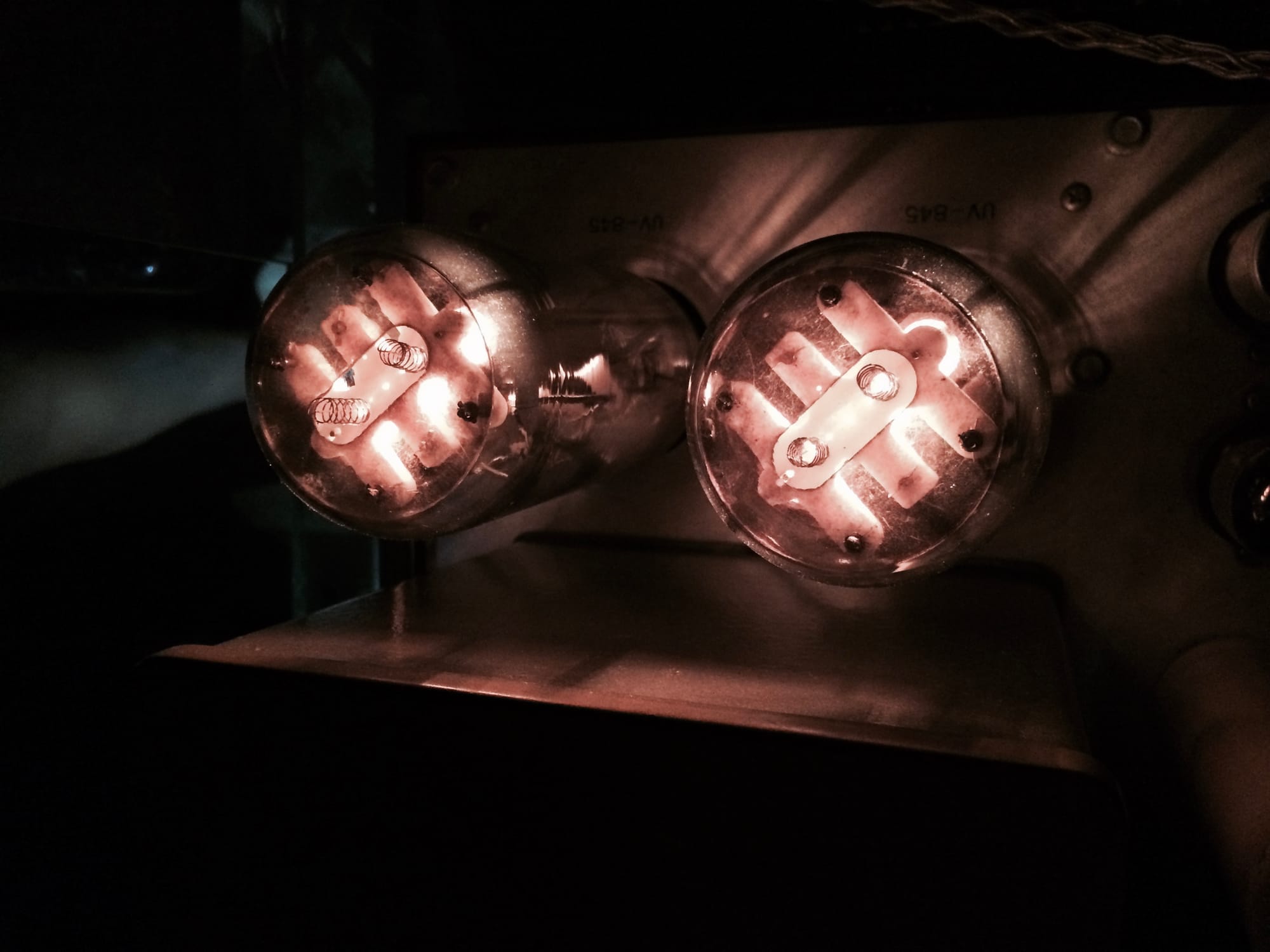 Two old-timey lightbulbs, side by side, attached to a metal box in the dark.