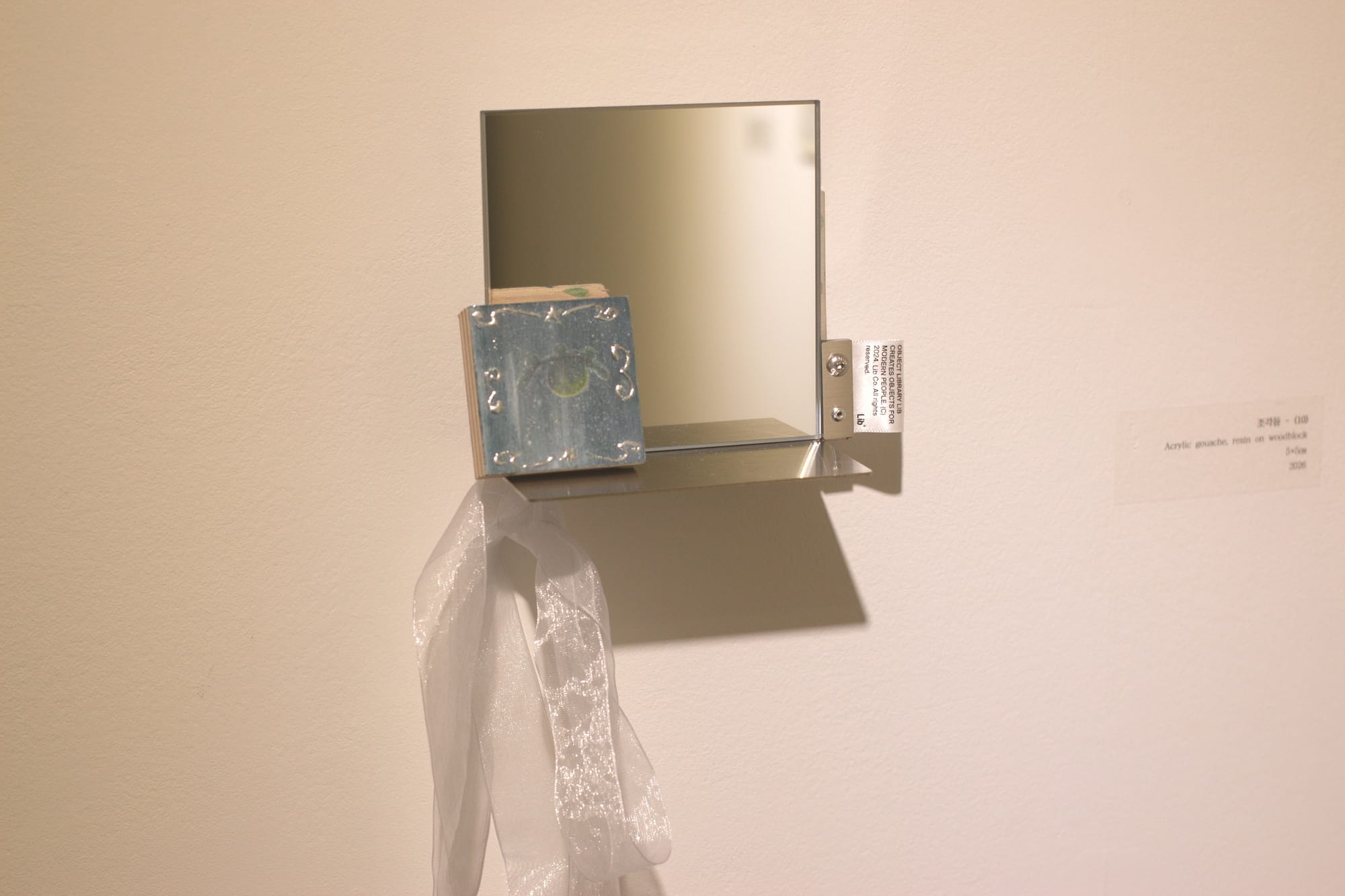 A small square mirror with a metal ledge and fixture with two screws, holding a fabric tag. On the ledge is an even smaller square blue-gray painting with shiny metallic 3D curlicues around the sides. There is an iridescent white ribbon attached.