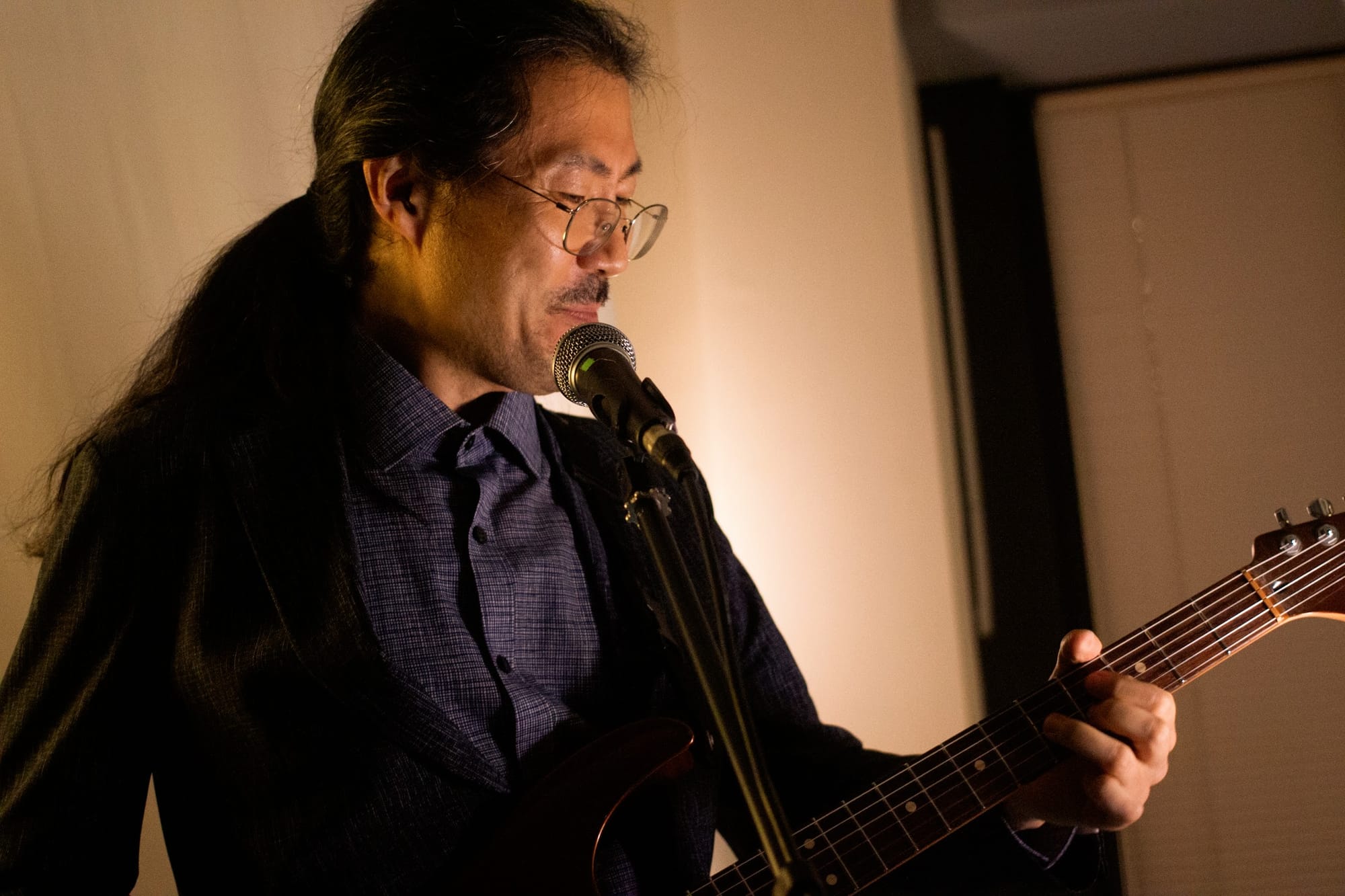 Hwang Chihyun, in glasses and a ponytail, smiles and plays electric guitar in front of a mic.