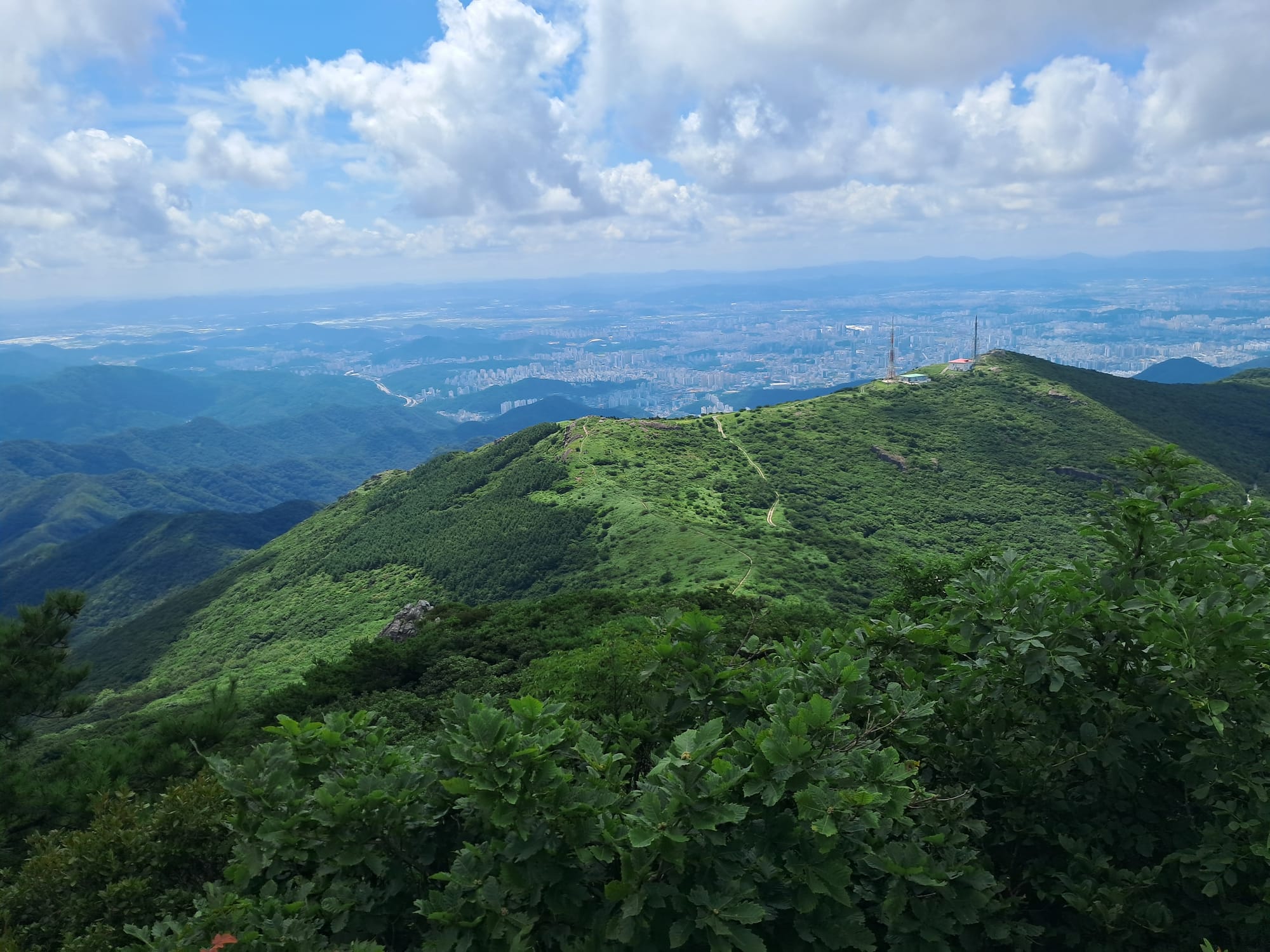 Clouds, rolling green mountains, and a city in the distance. There is a thin path visible along one of the mountains.