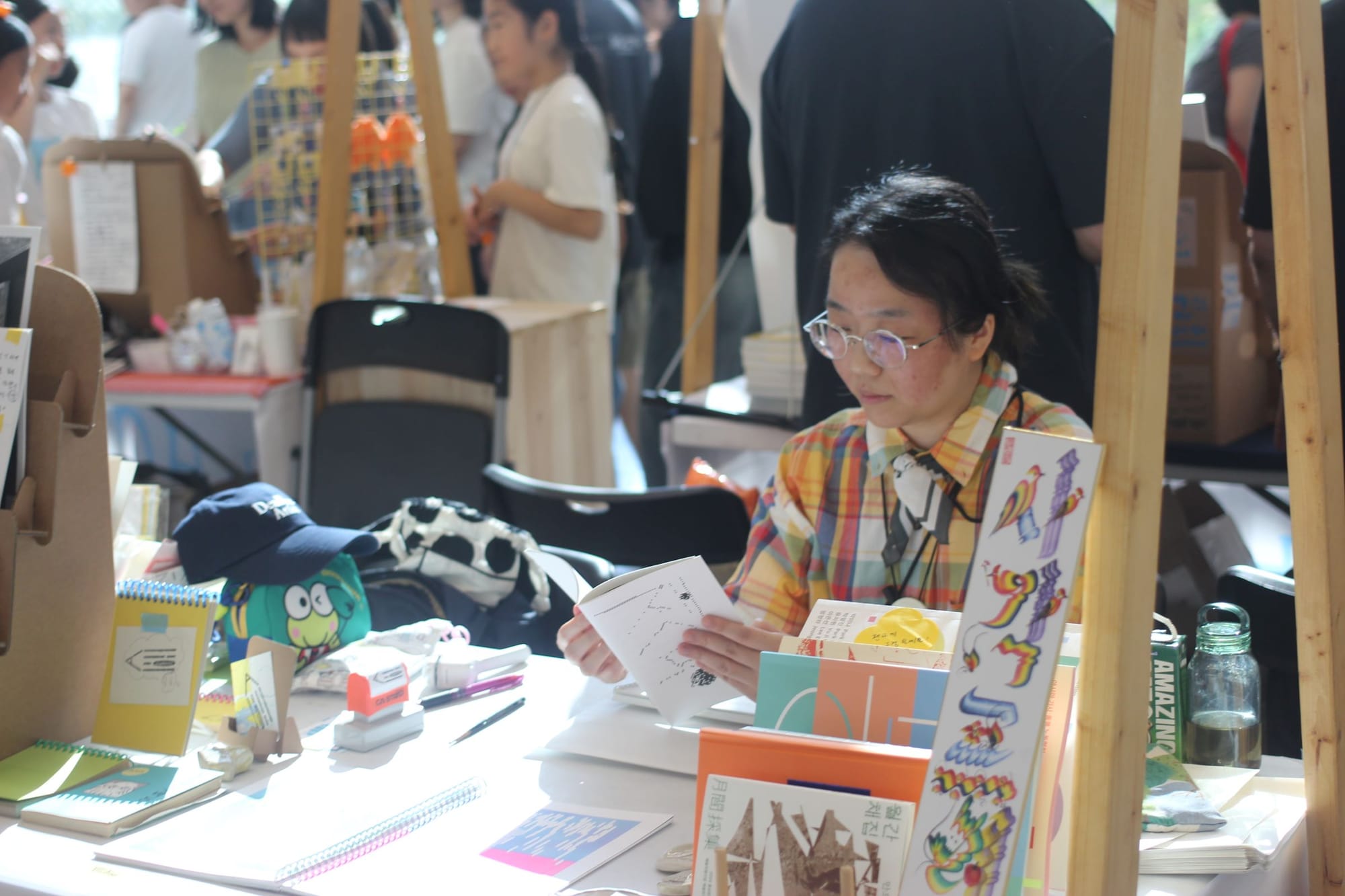 Jisun Choi, in a yellow plaid shirt, reads a book while sitting at her booth. The table in front of her has colorful, cute illustrations and prints. A sign says Sagak Press in Korean.