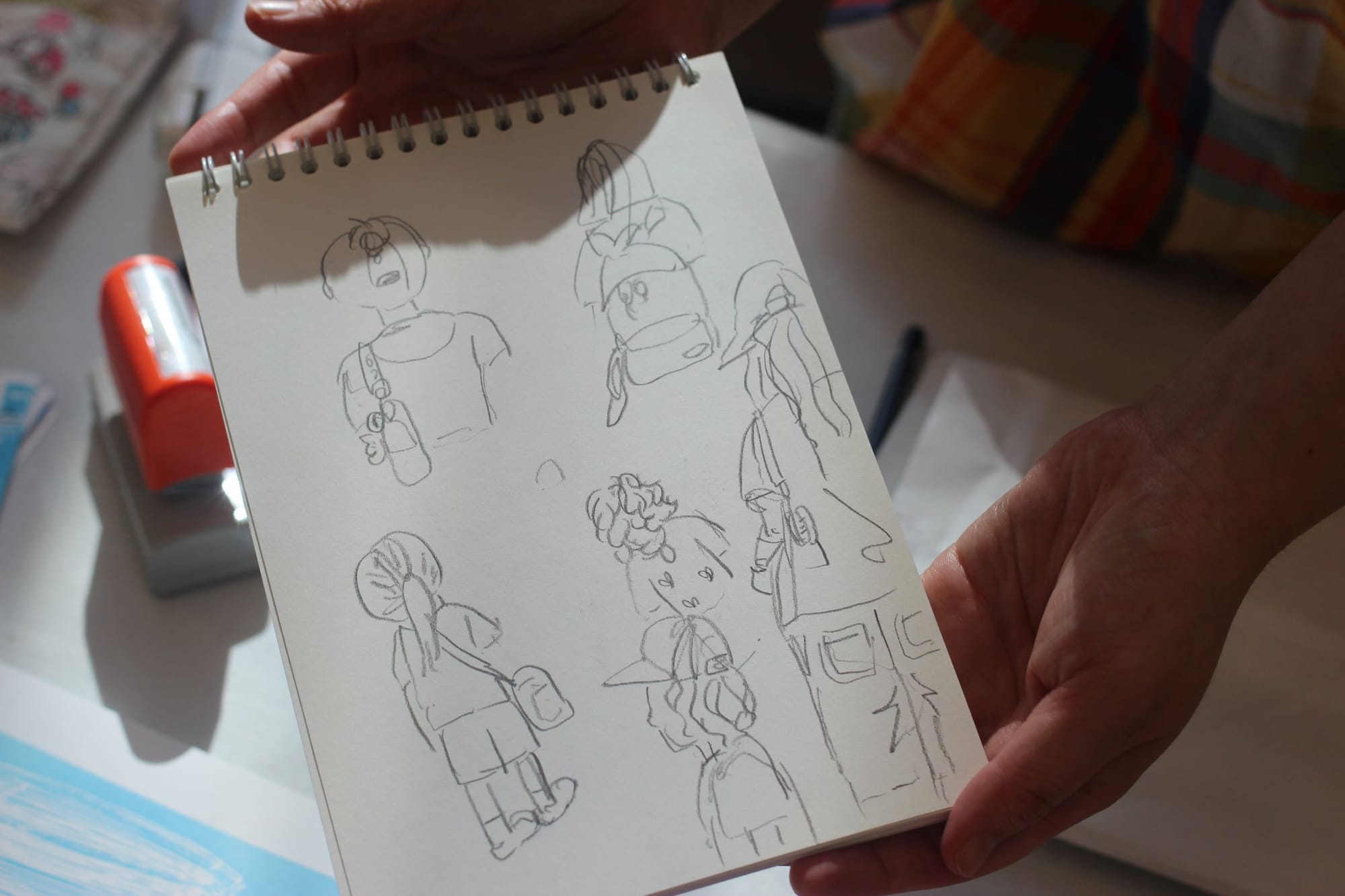 Two hands hold a sketchbook with several small sketches of passersby.