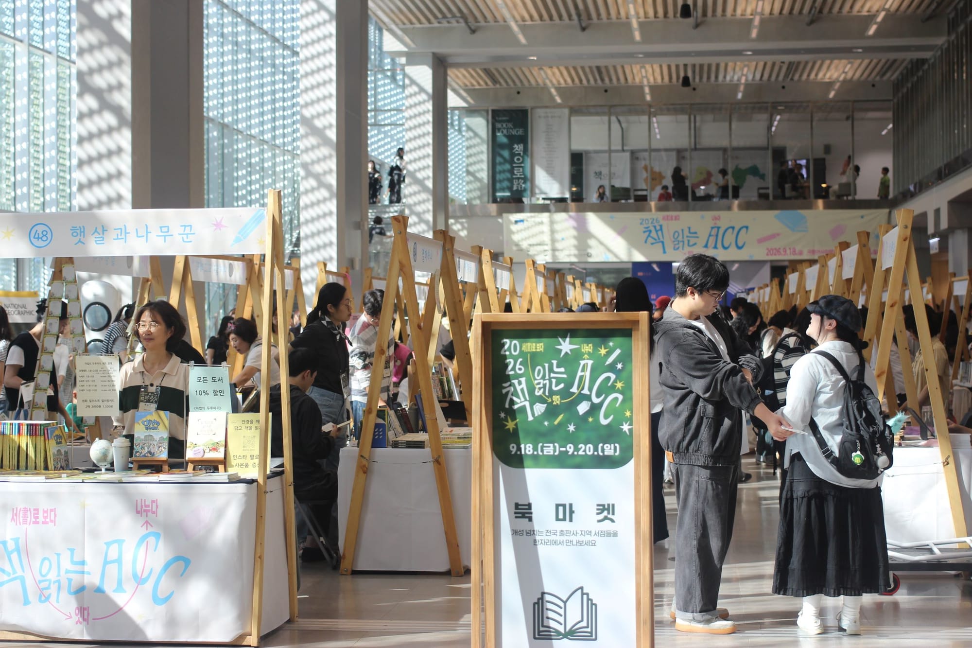 Three long rows of booths in a large sunlit space, with plenty of people walking through the aisles and manning the booths.