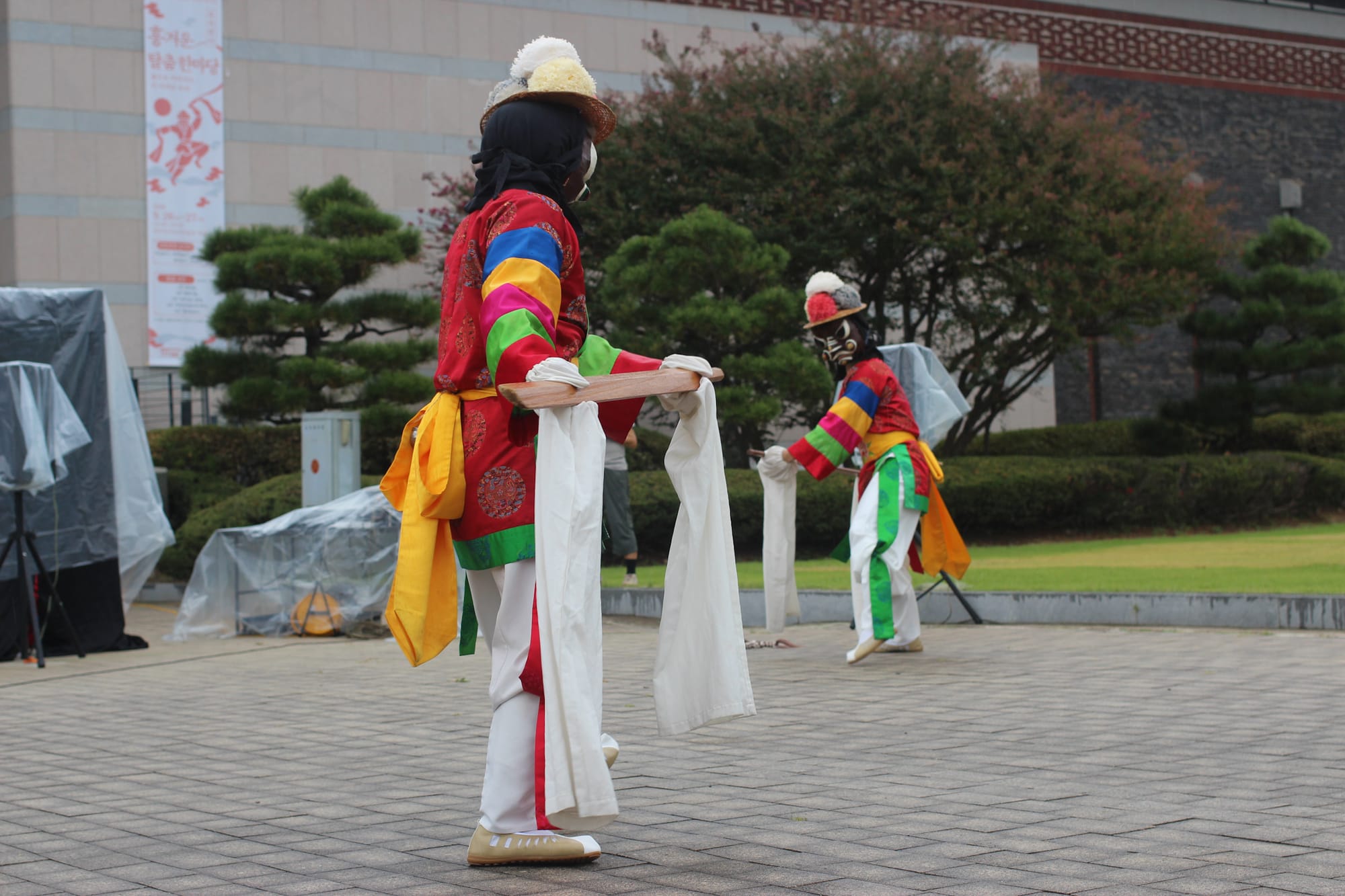 Two dancers with staffs face each other on a museum pavilion. They are wearing masks with white facial features and hats with round puffballs.