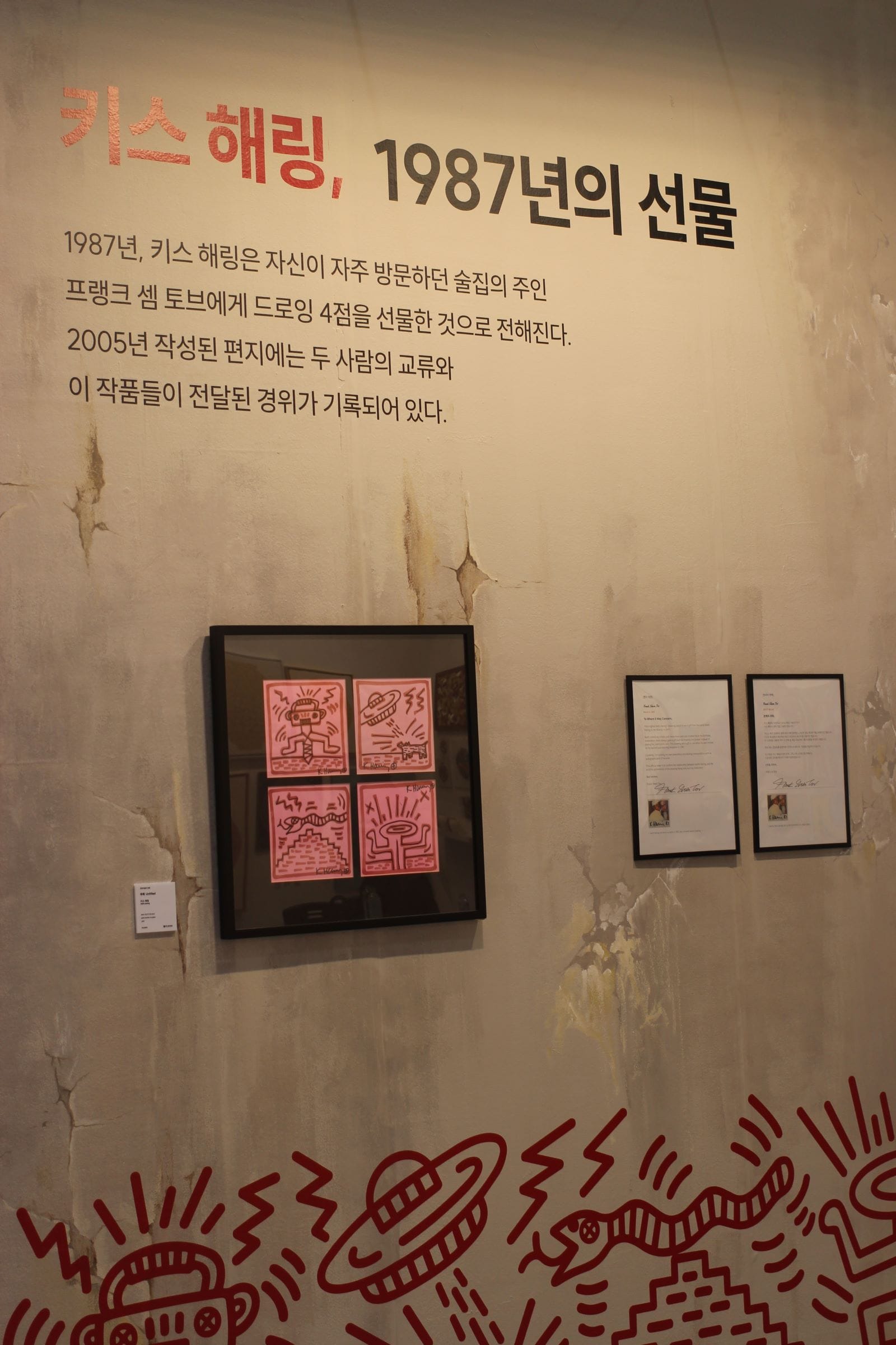 A wall that appears cracked and aged, with a framed four part drawing series of iconographic red images on pink paper.