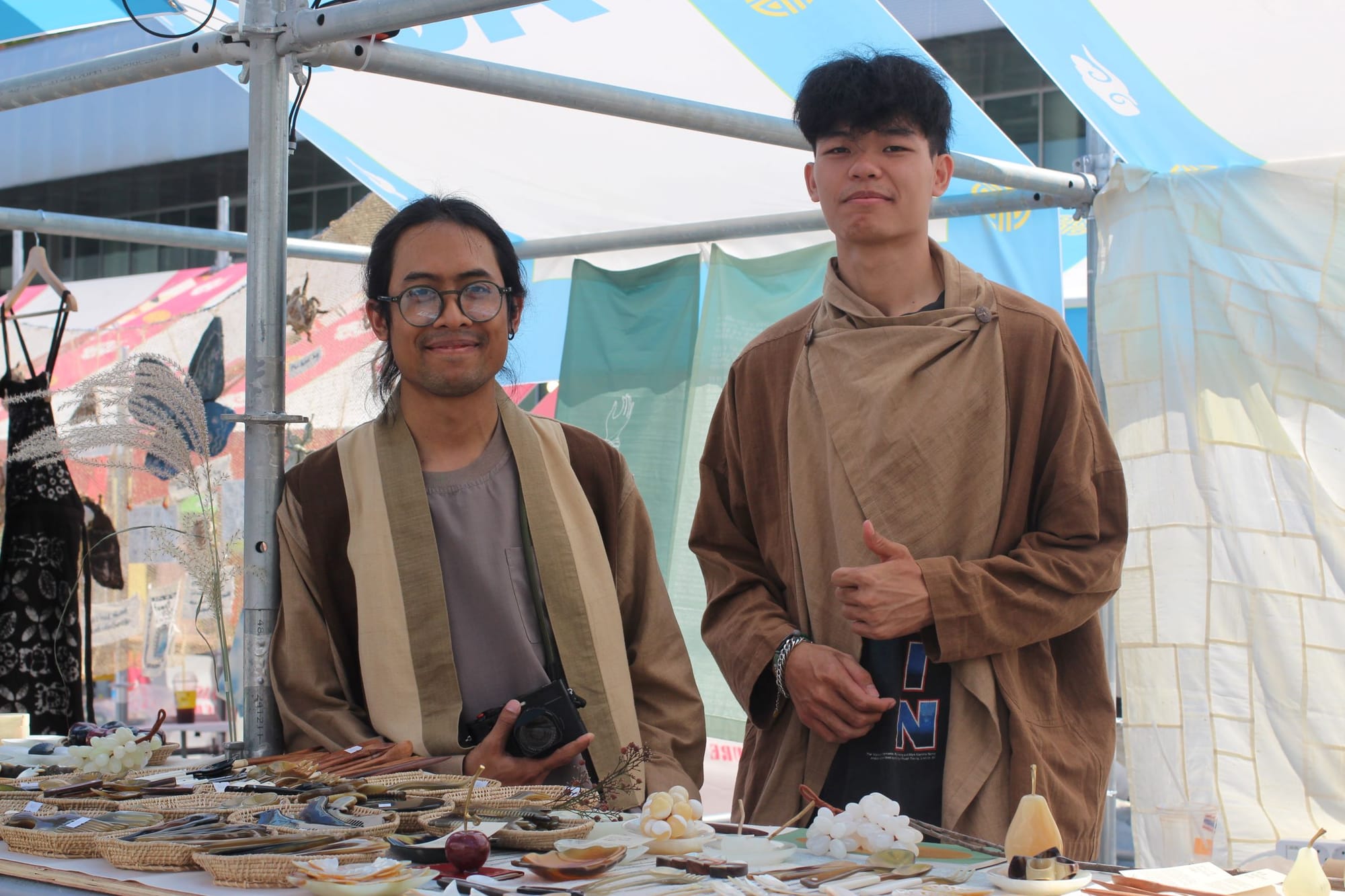Kittikhun and Tree stand at their stall of artisan accessories and small objects, including stone grapes. In the back are draped fabrics.