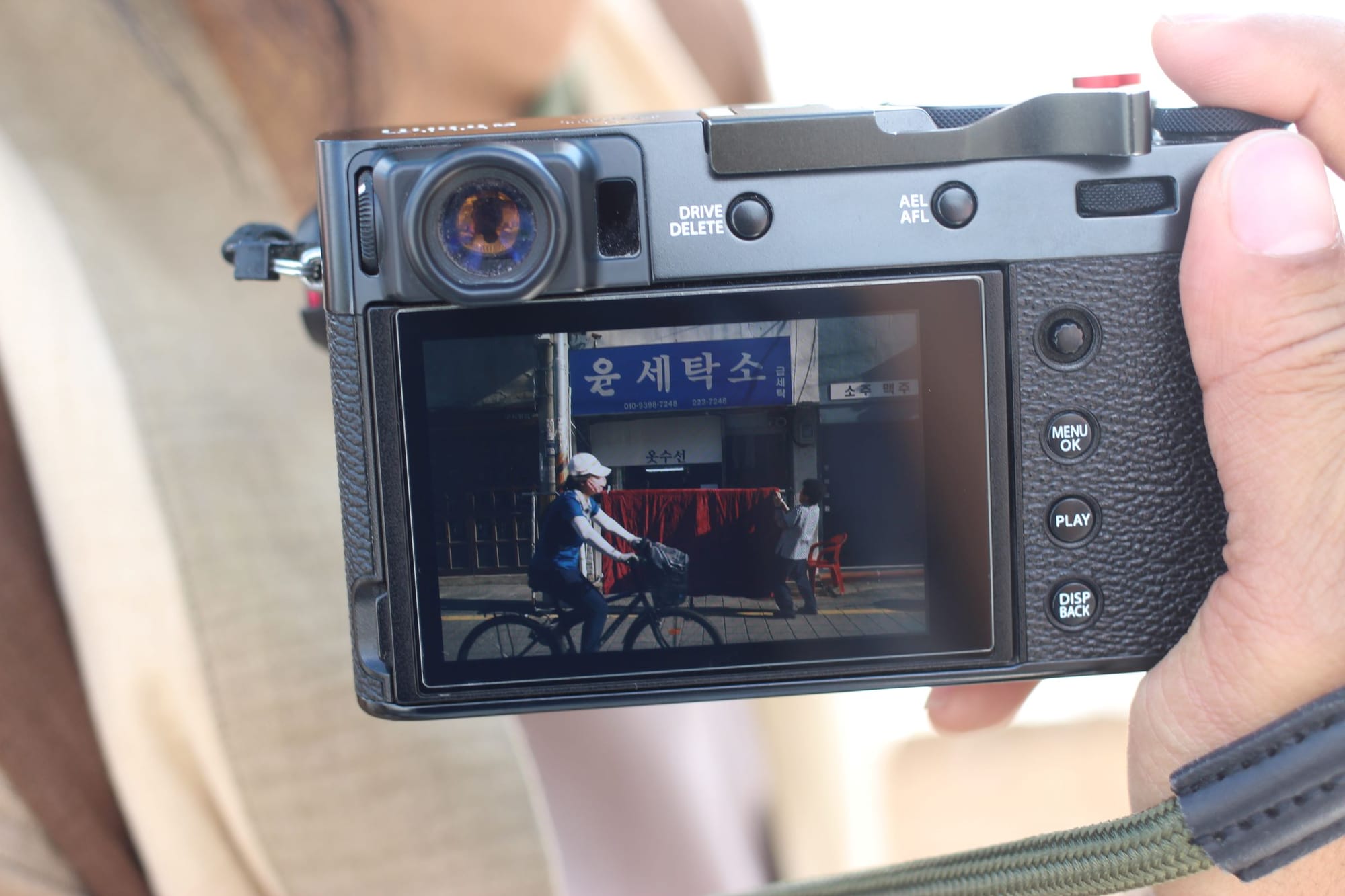 A hand holds a camera showing a photo taken in Korea of a person on a bike riding past a blue sign.