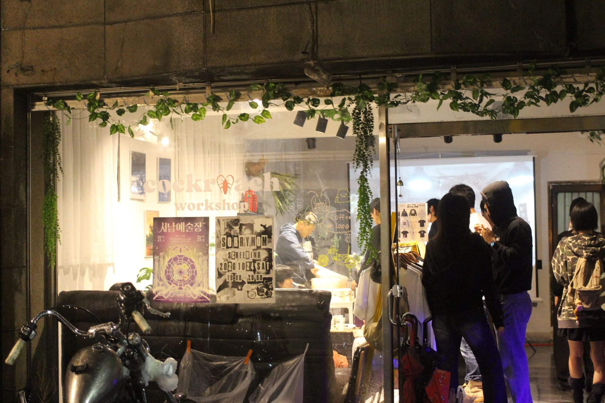 Exterior of the workshop. The walls are concrete, with an inset glass section. There is a cockroach logo on the glass and two posters, one for the SORRYMOM screening. About ten people mingle.