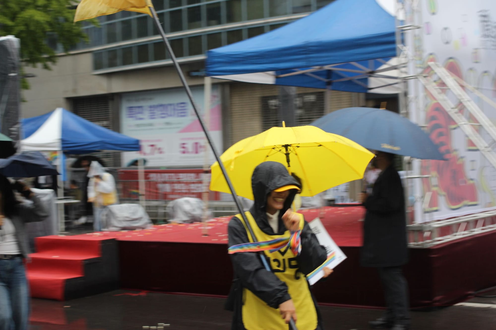 A person wearing yellow, with a flag on a tall stick, smiles in the rain, slightly blurry.