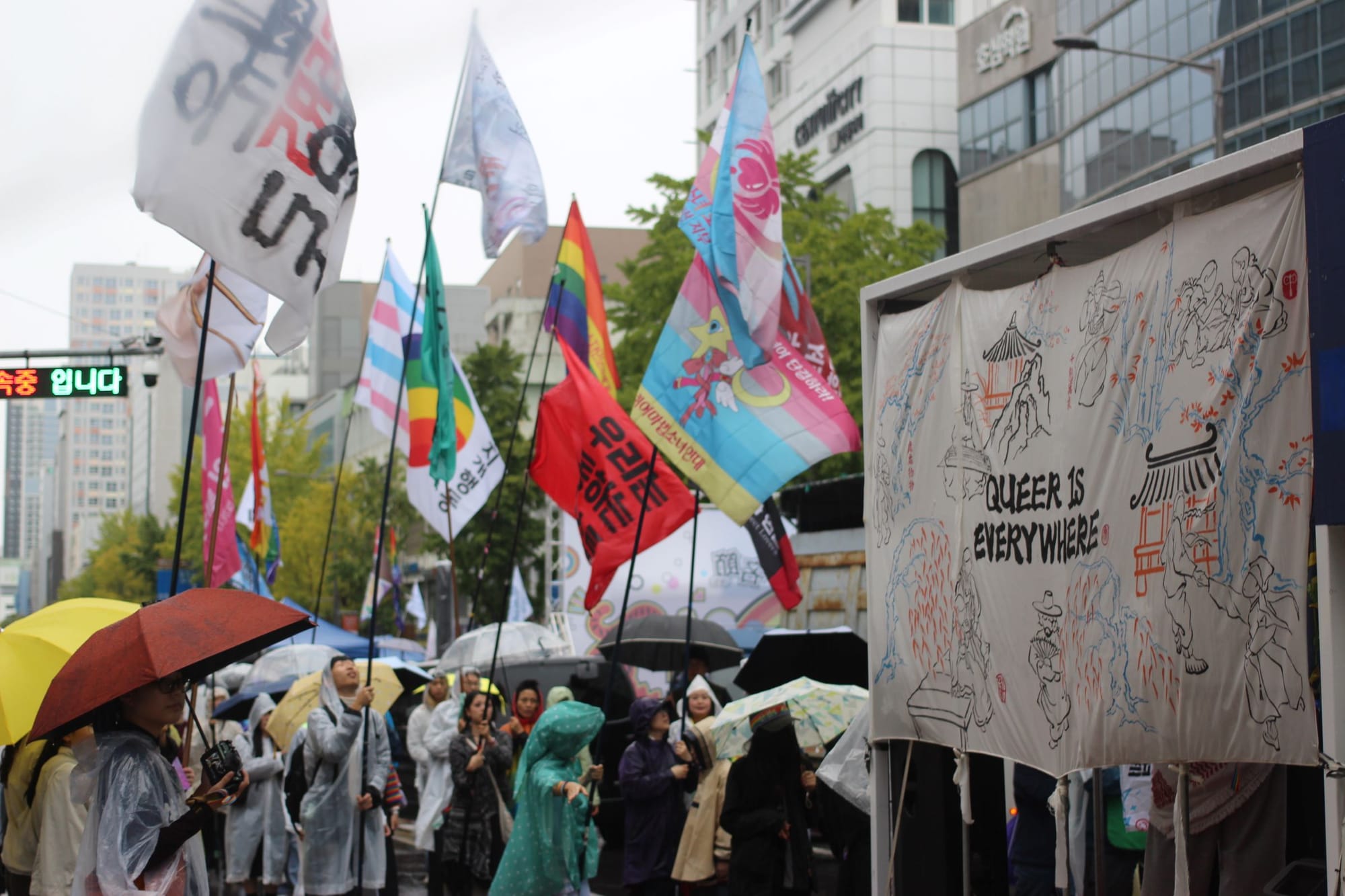 Many colorful flags on long sticks, held by people with umbrellas. A large banner says "QUEER IS EVERYWHERE."