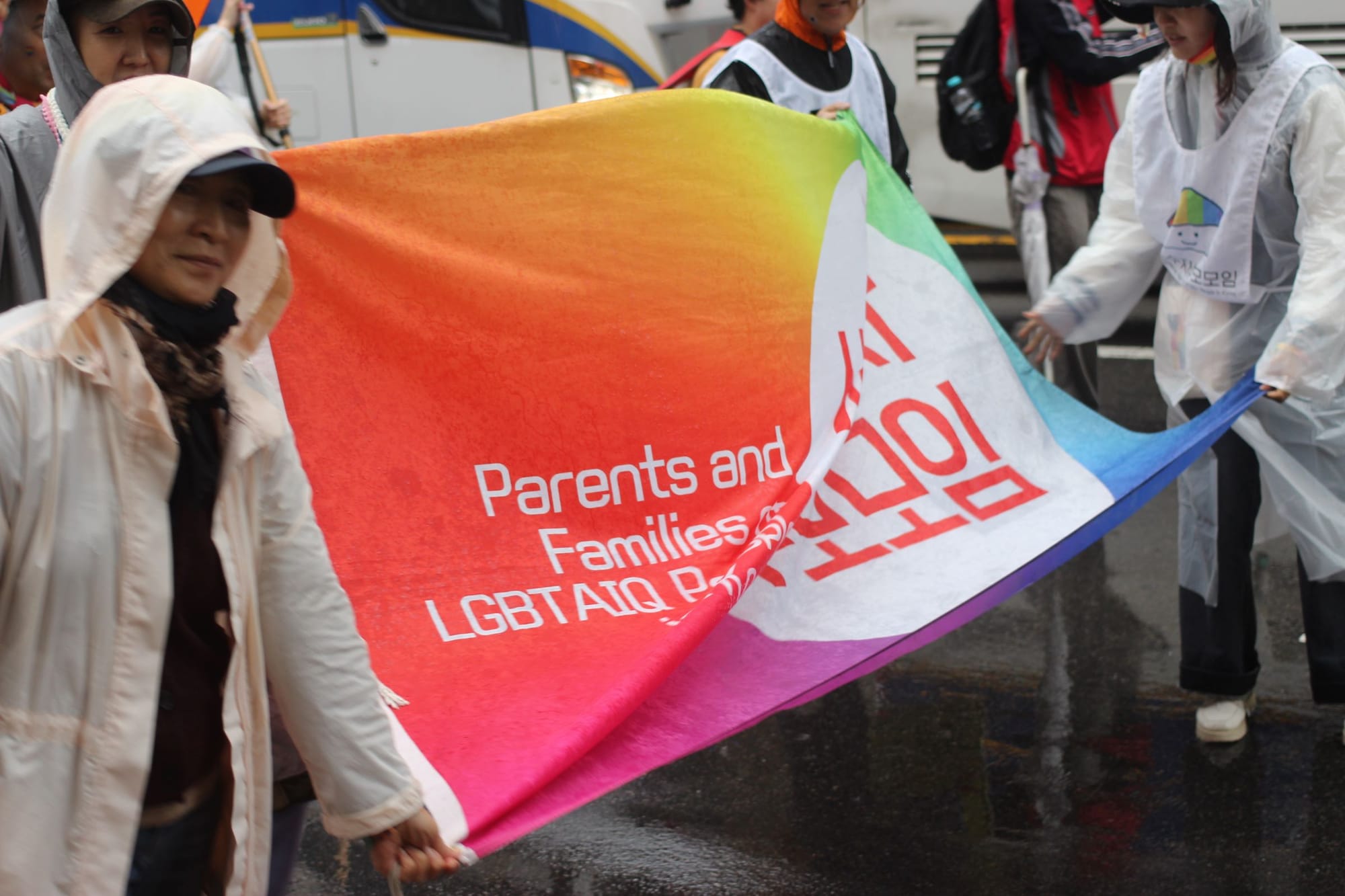 In the rain, a handful of people hold a large rainbow banner. You can make out the words "Parents and Families" and "LGBTAIQ."