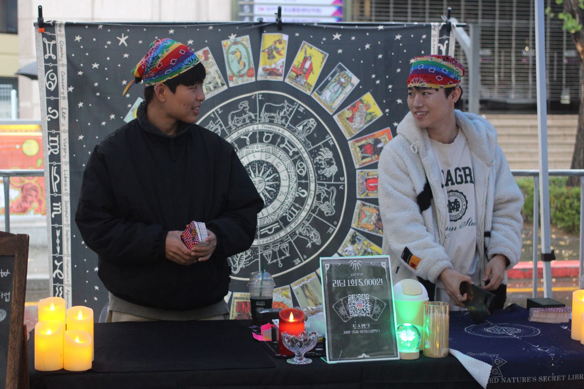 Two people with rainbow bandanas hold card decks and look toward each other, at a booth with fake candles and an occult zodiac and tarot backdrop.