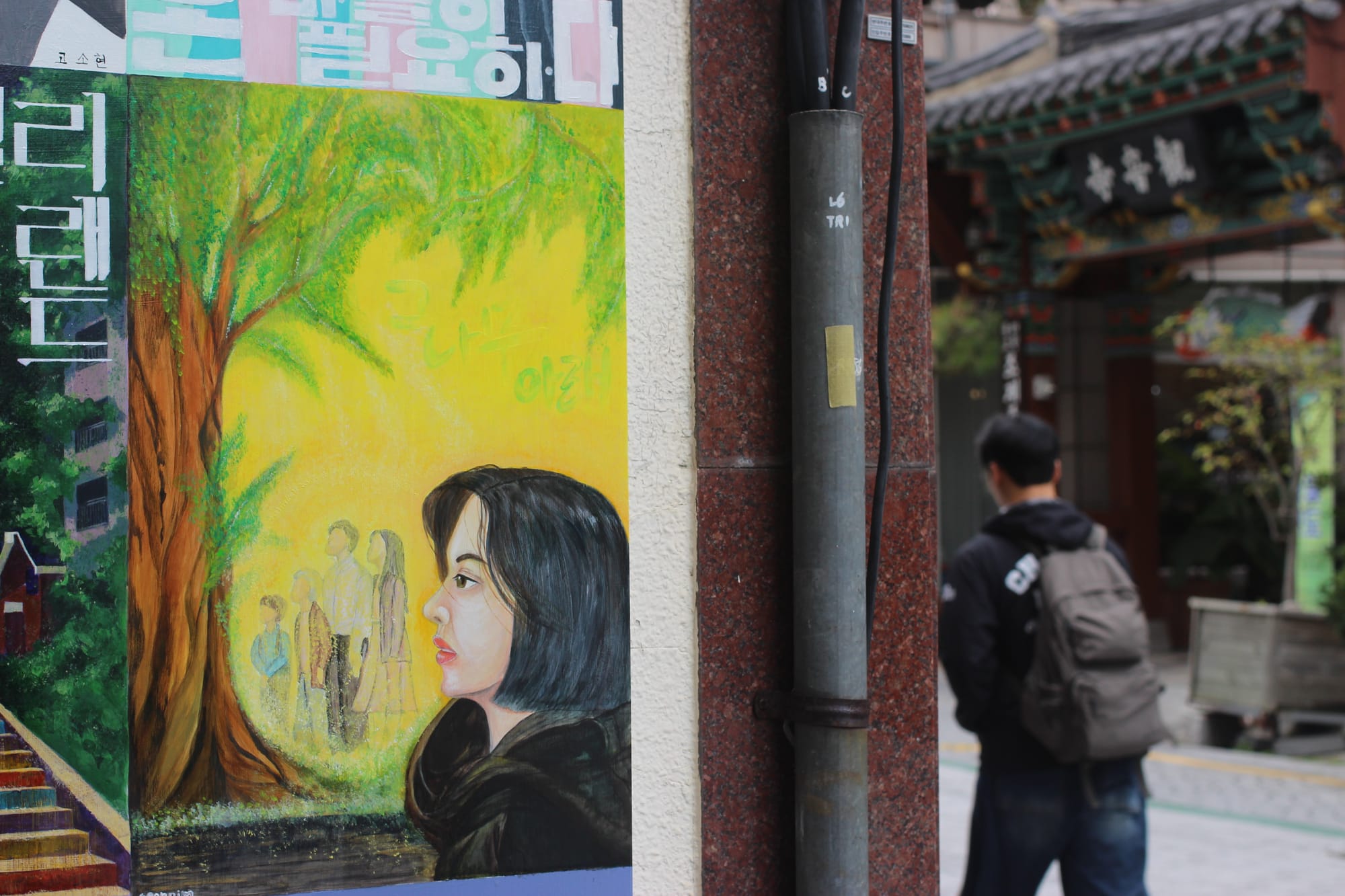 A painting with a large tree with green leaves against a yellow background. There are hazy figures, young and old, underneath. In the foreground is a woman looking in the direction of the tree. Past the painting is a passerby with a backpack.