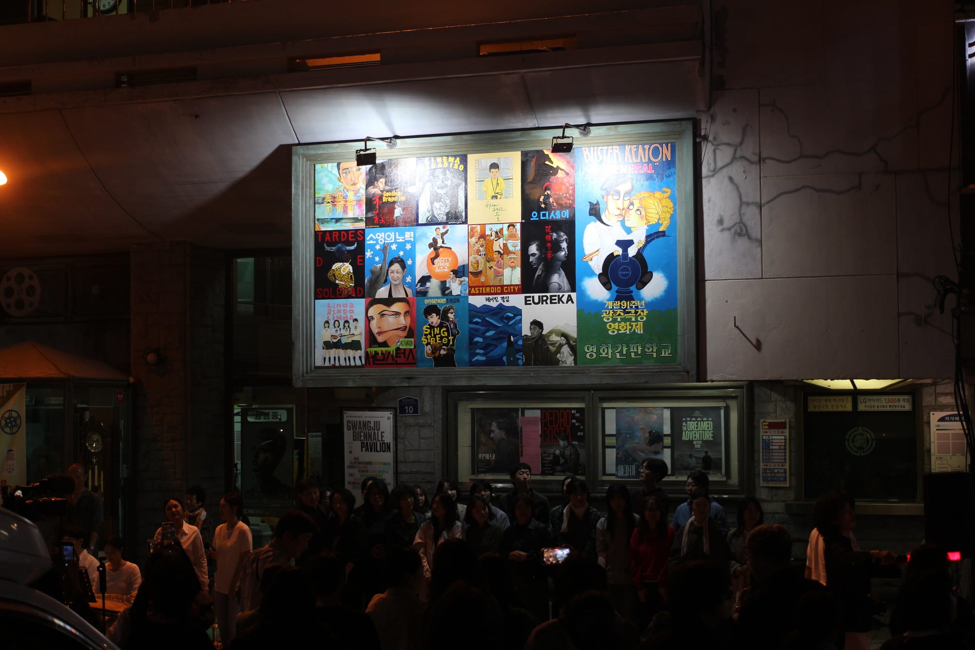The movie signboard at night, illuminated by two spotlights. Underneath, in the dark, is a crowd of people.