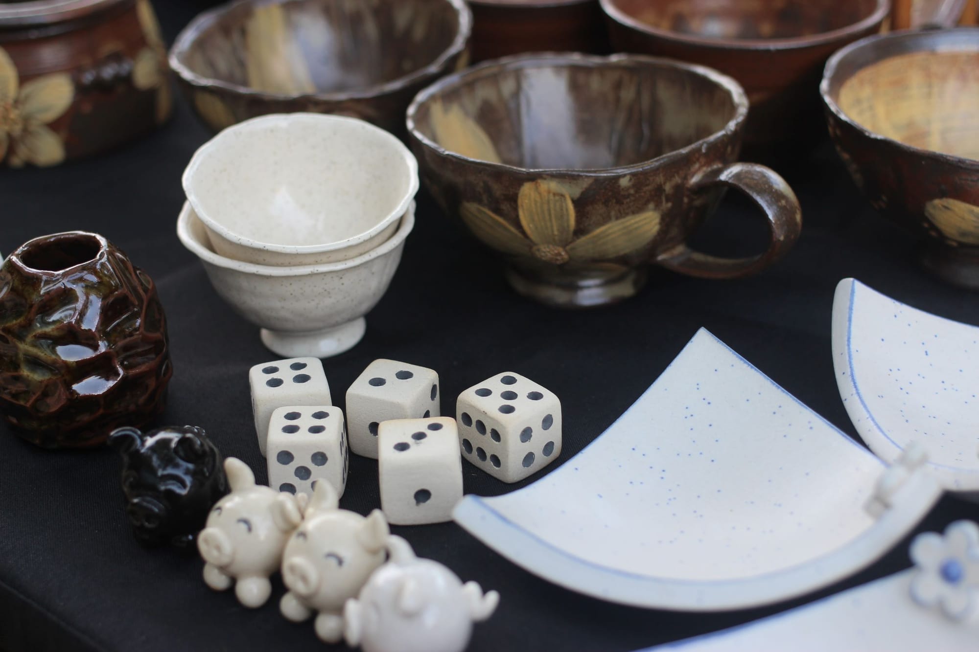 A display of ceramics on a table. There are tiny ceramic pigs, simple cups, blue-rimmed white pieces arranged to look like a flower, and ceramic dice.
