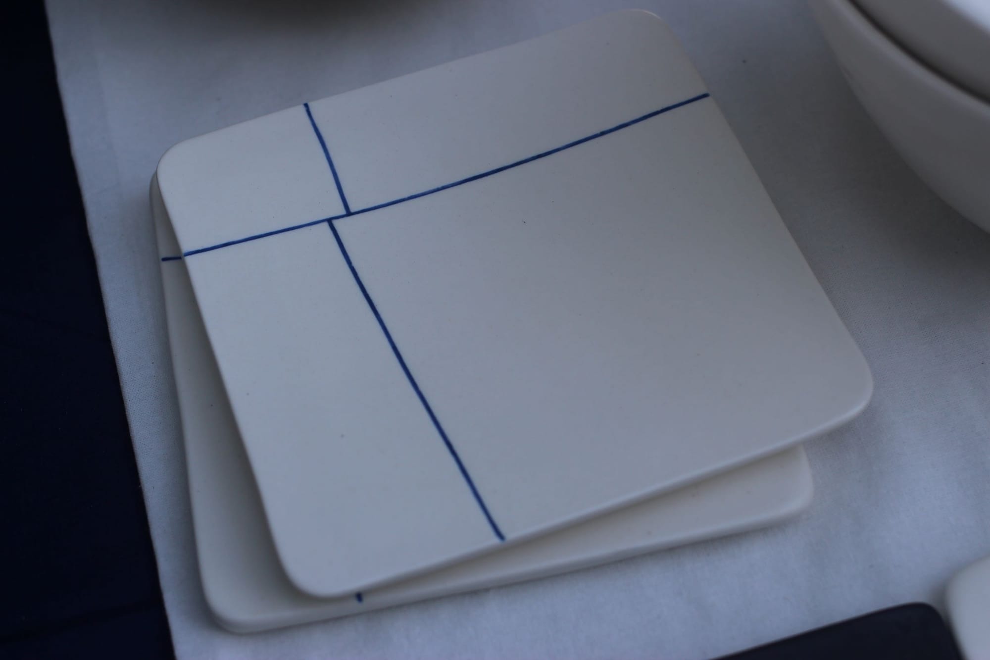 Two ceramics coasters. They are white squares with rounded edges and narrow, dark blue lines crossing through at almost-right angles.