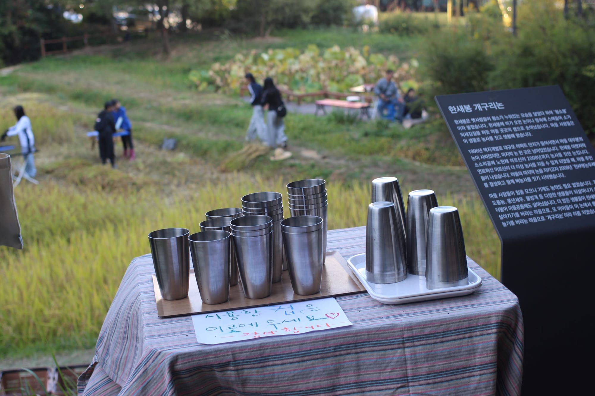 Shiny metal cups on a table. Behind, out of focus, are people working and walking in a green field.