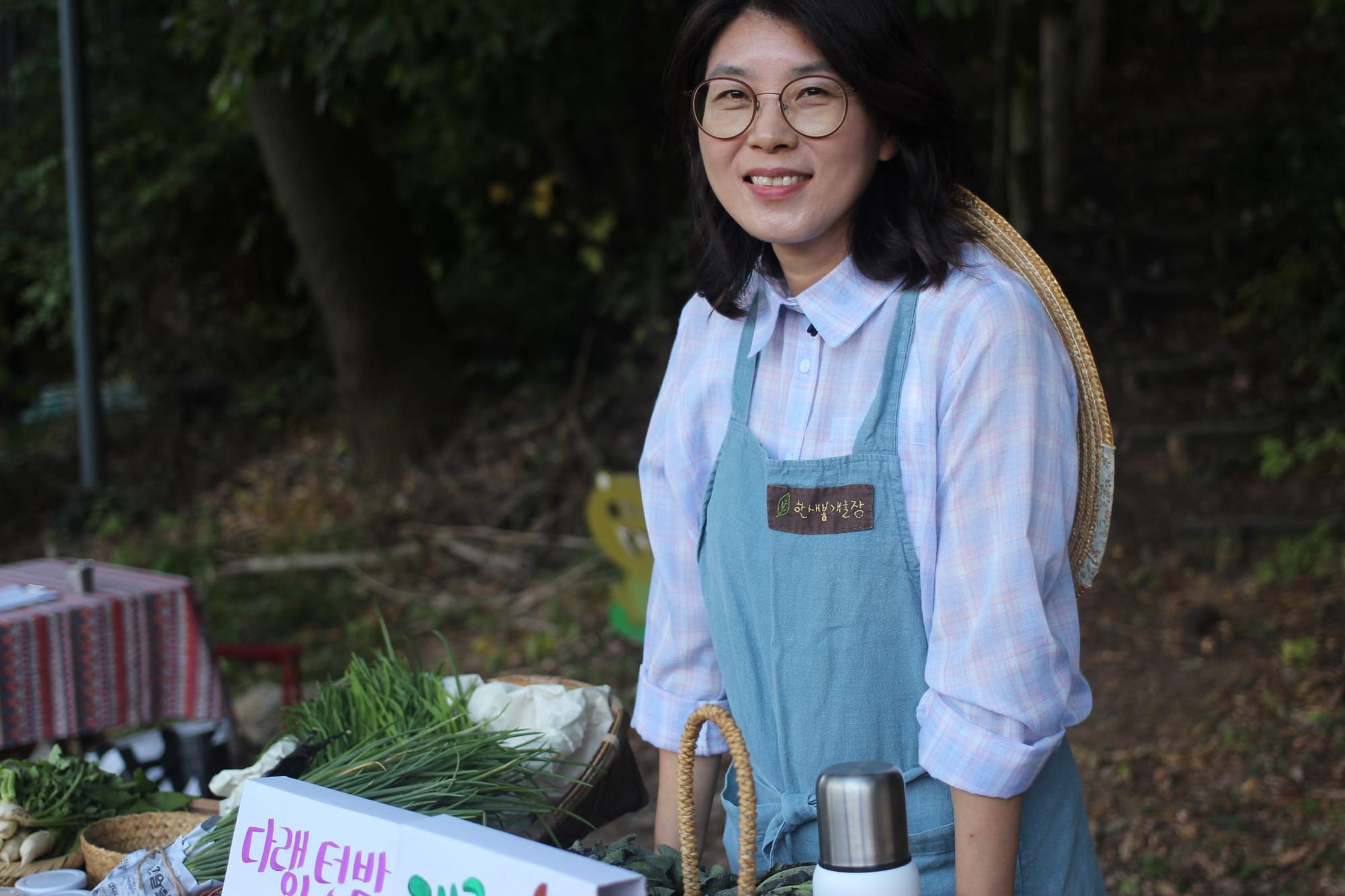 Kim Seonim smiles, wearing a light blue apron. Her farmer's hat rests on her back. Next to her are green onions and radishes.