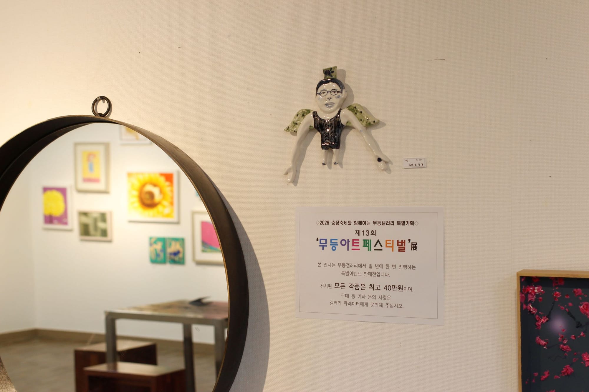 The ceramic man, with green wings and a green cup above his head, is now near a circular mirror and a different framed piece. The mirror reflects several vibrant paintings on the opposite wall.