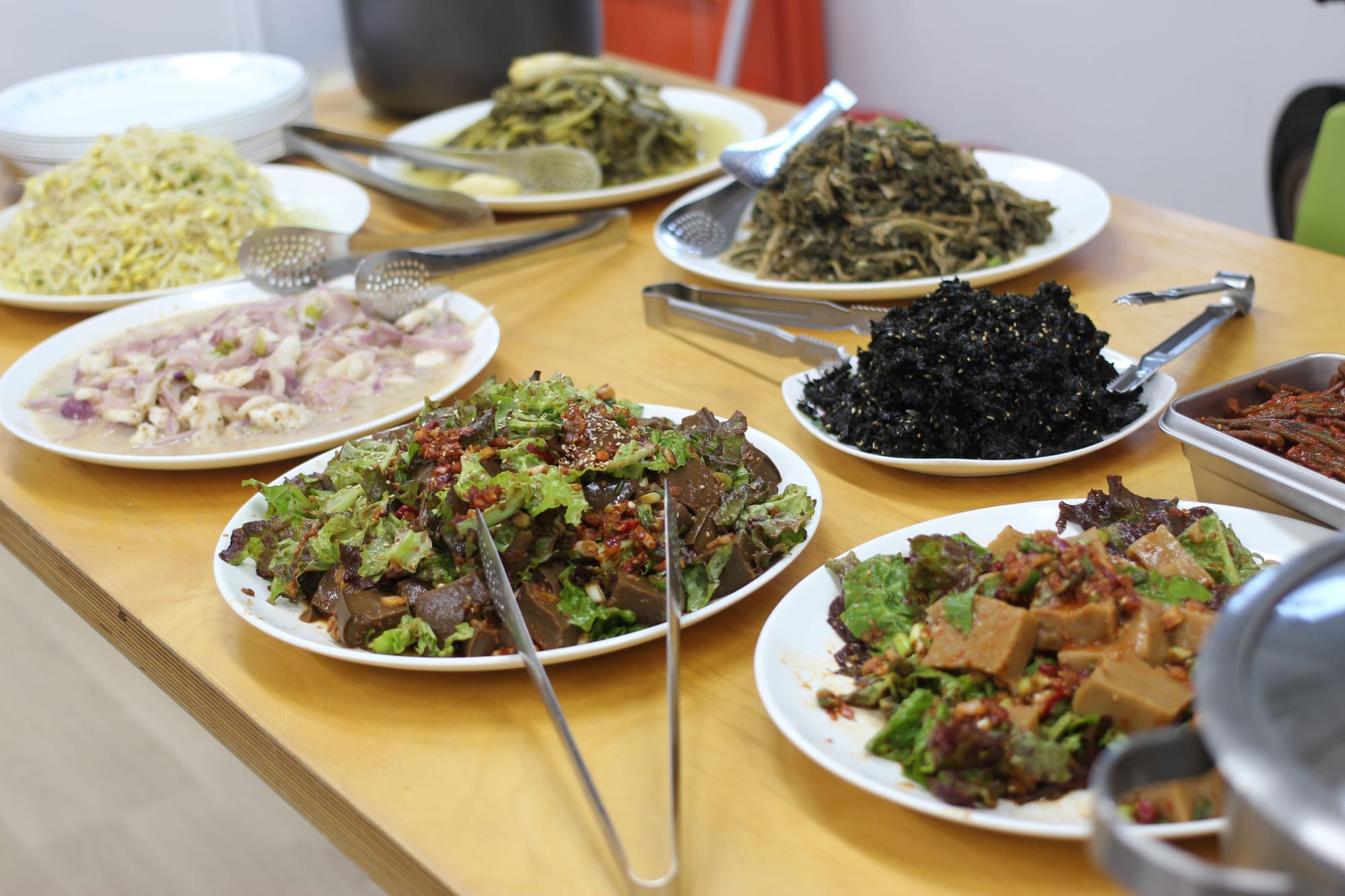 A table of several plates piled high with different vegetable dishes: fried seaweed, seasoned lettuce and muk, beansprouts, onions.
