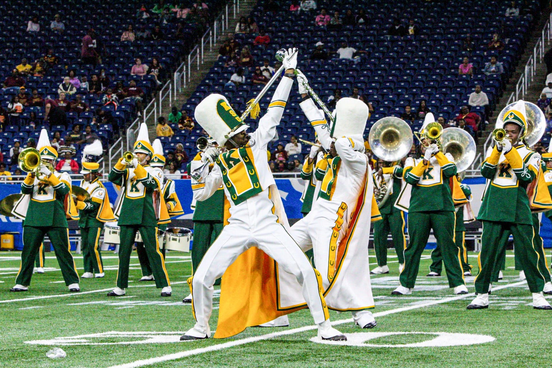 A marching band performs on a football field, with two members in white uniforms crossing their batons at the front while others in green and yellow play brass instruments. Photo credit: Akee