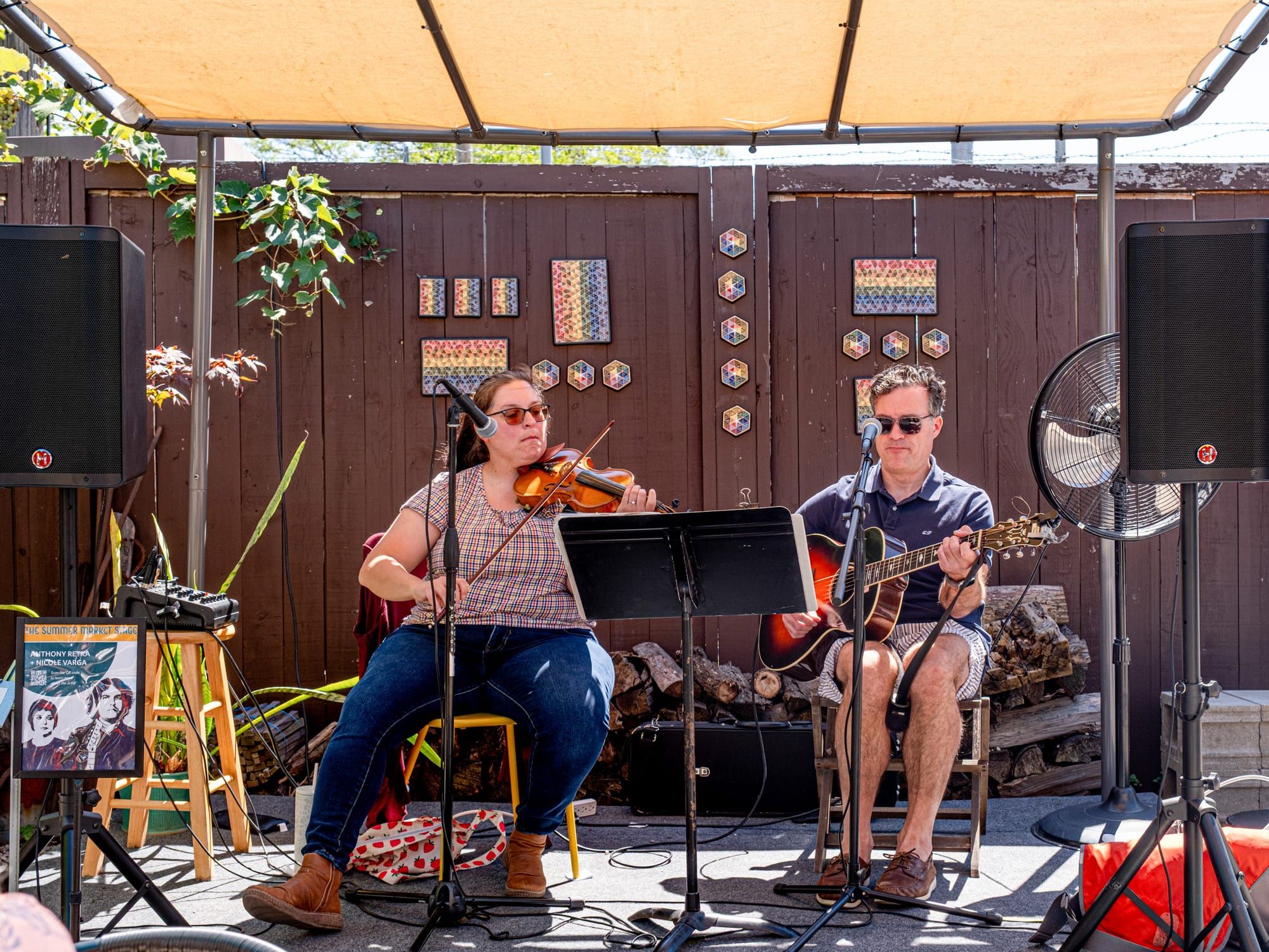 Two musicians sit on a stage under a tent, one playing the violin and the other playing the guitar.